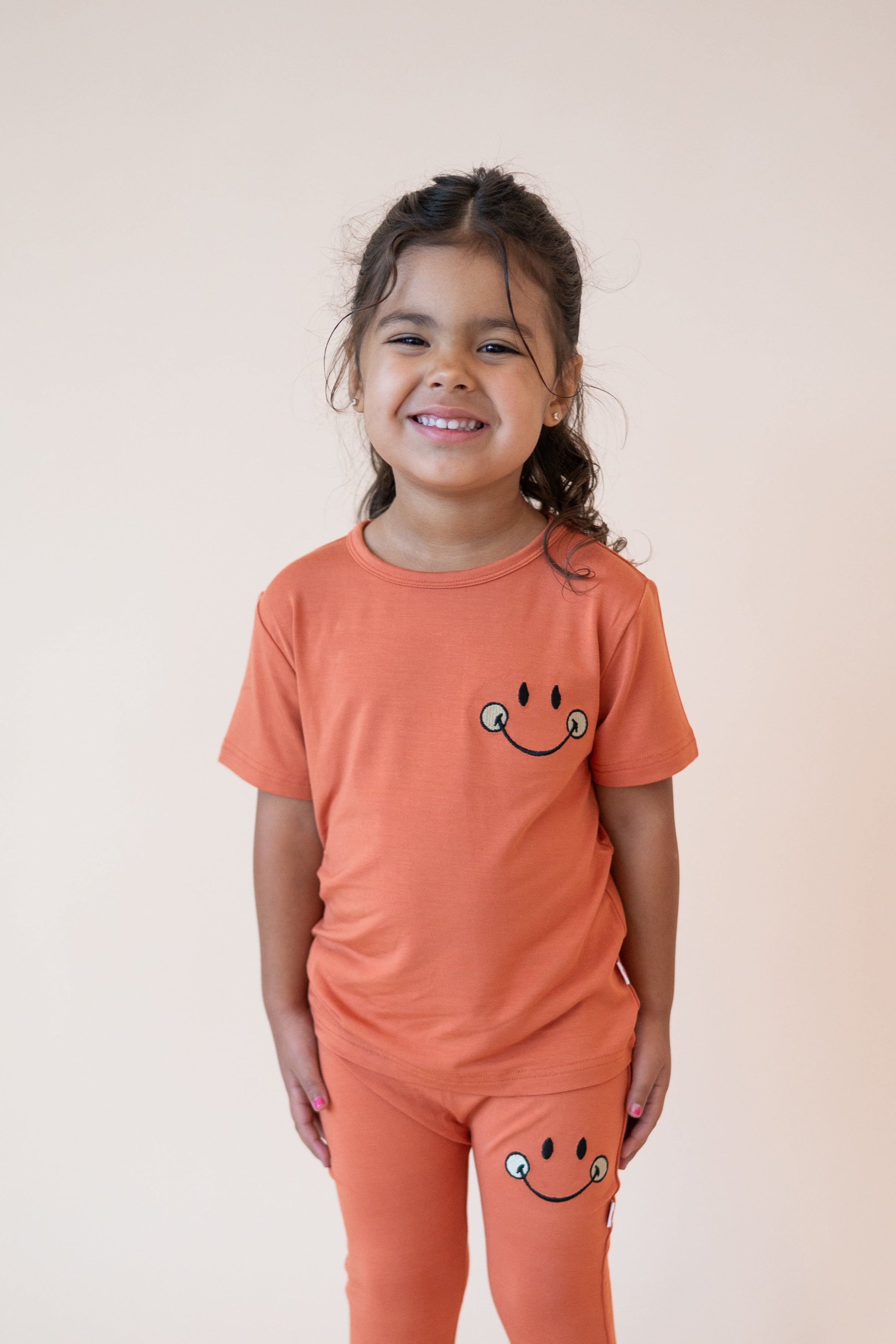 A young girl smiles in an orange t-shirt and pants with a smiley face design.