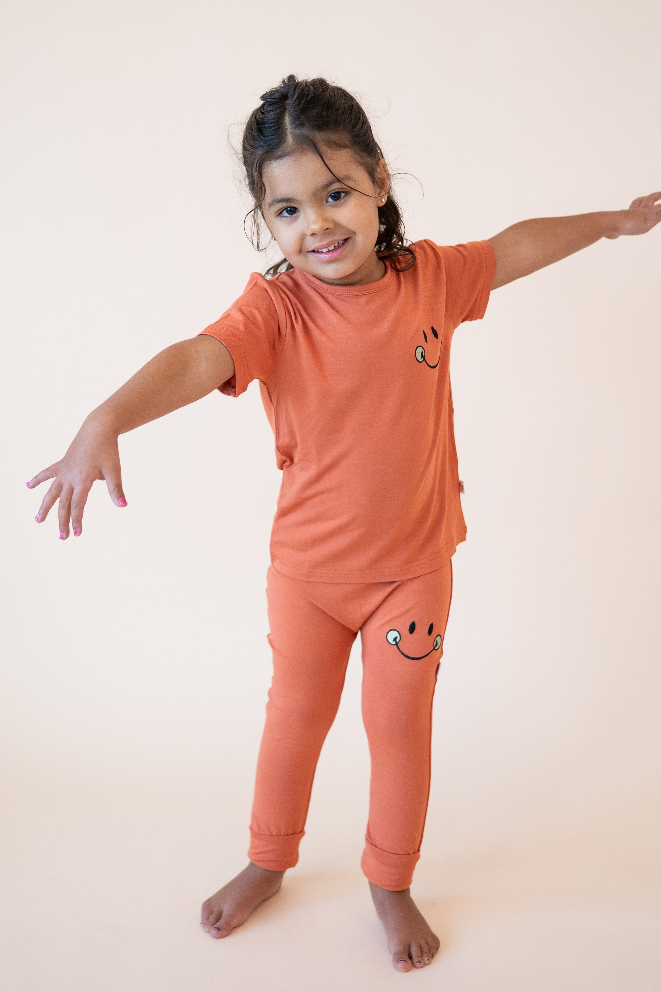 A young girl wears an orange t-shirt and pants with smiley face designs.