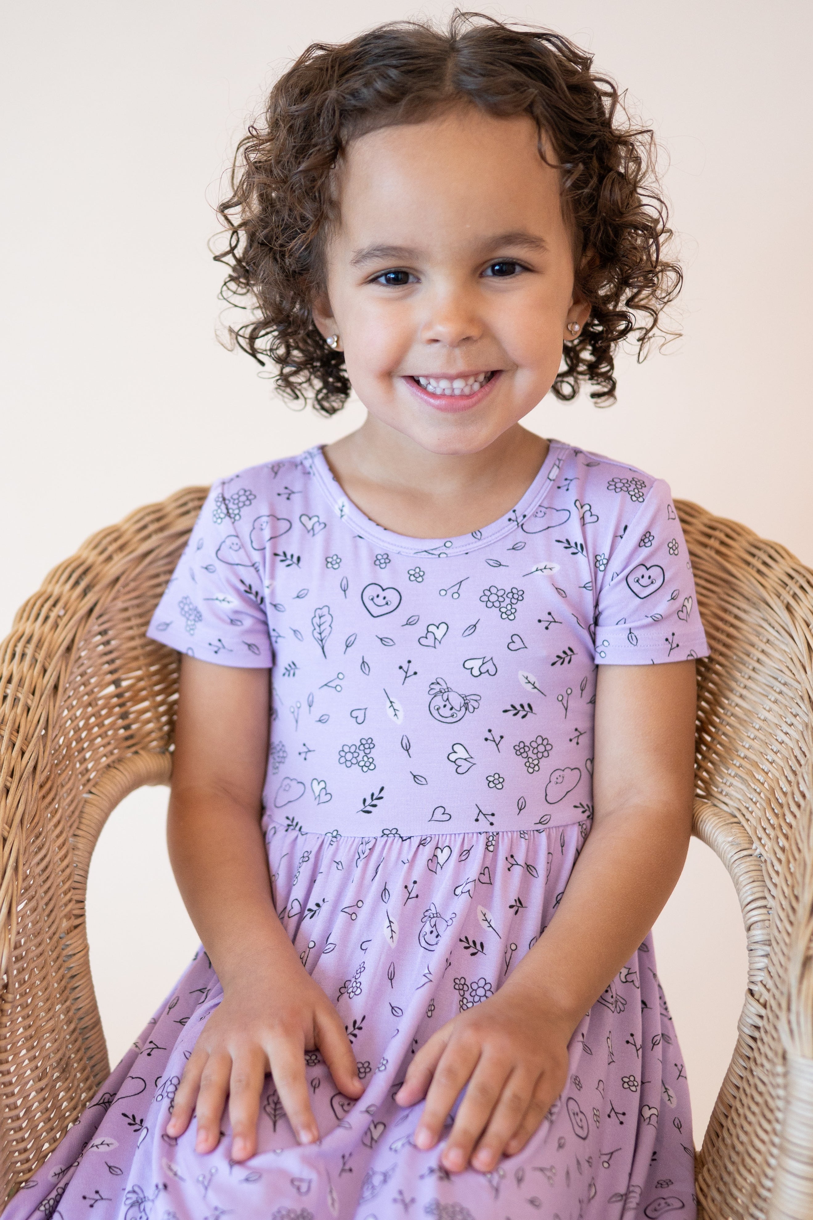 A young girl smiles while sitting in a wicker chair wearing a lilac dress with a doodle print.