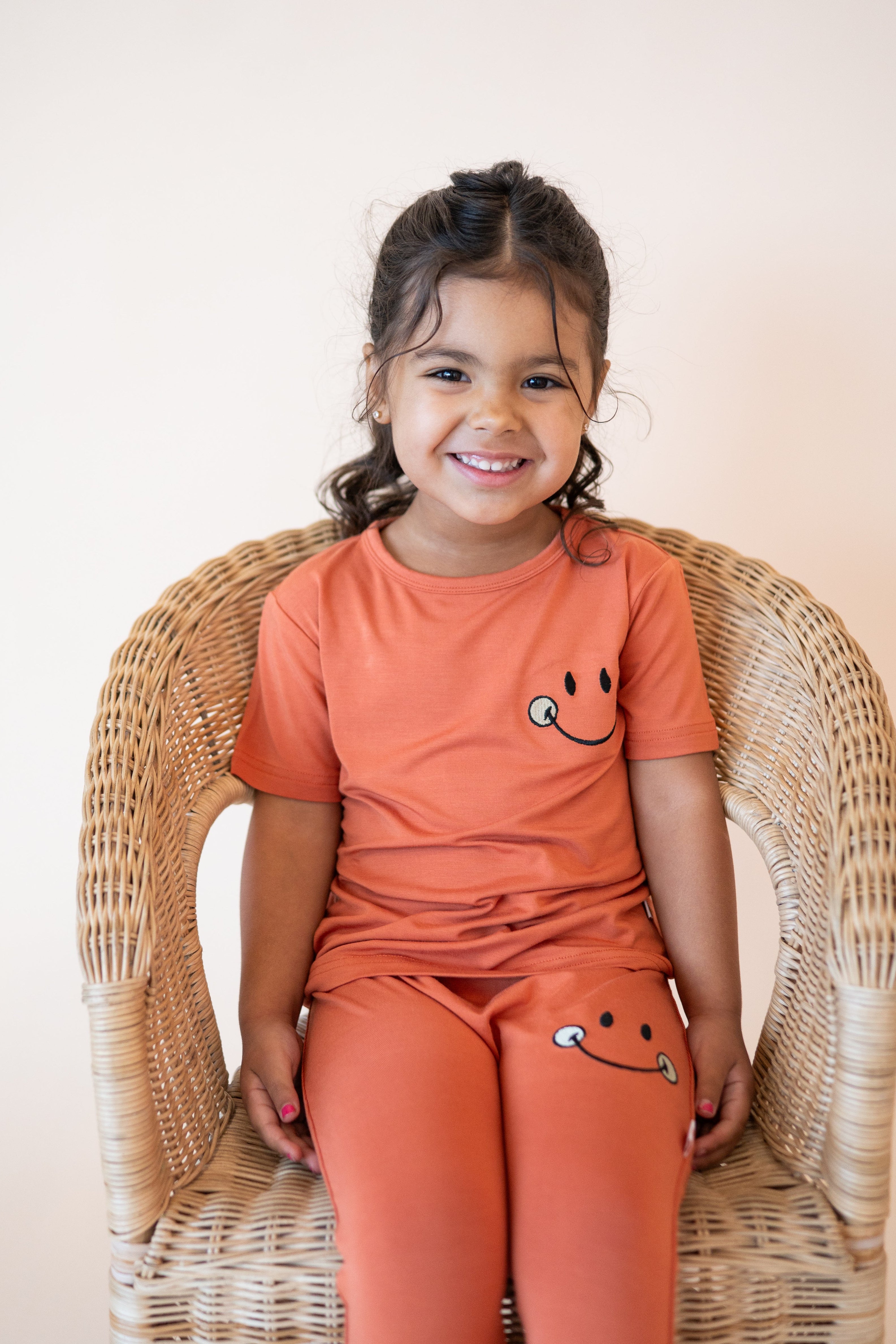 A young girl in a rust-colored t-shirt with a smiley face sits in a wicker chair.