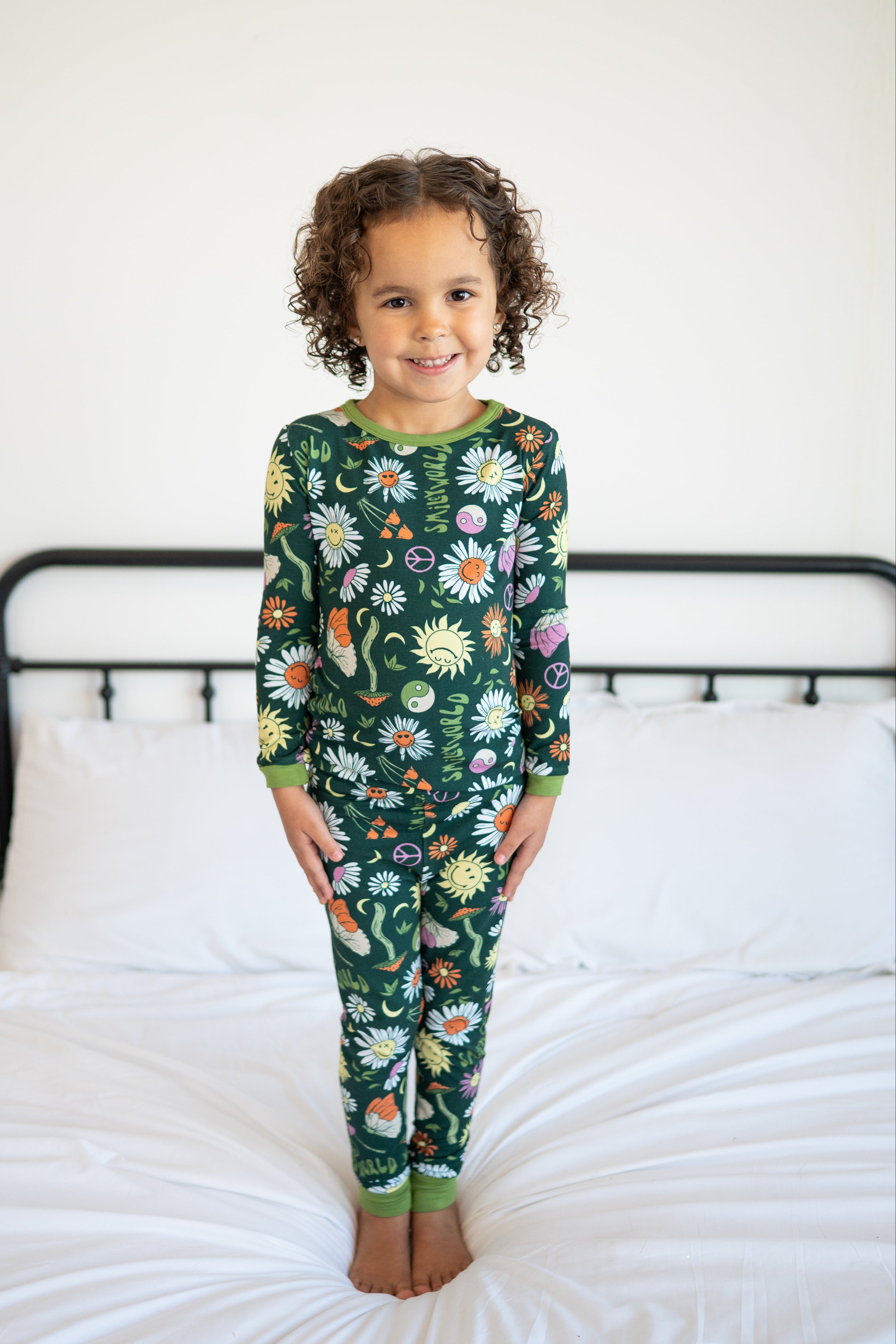 A young girl stands on a bed wearing green pajamas with a smiley flower print.