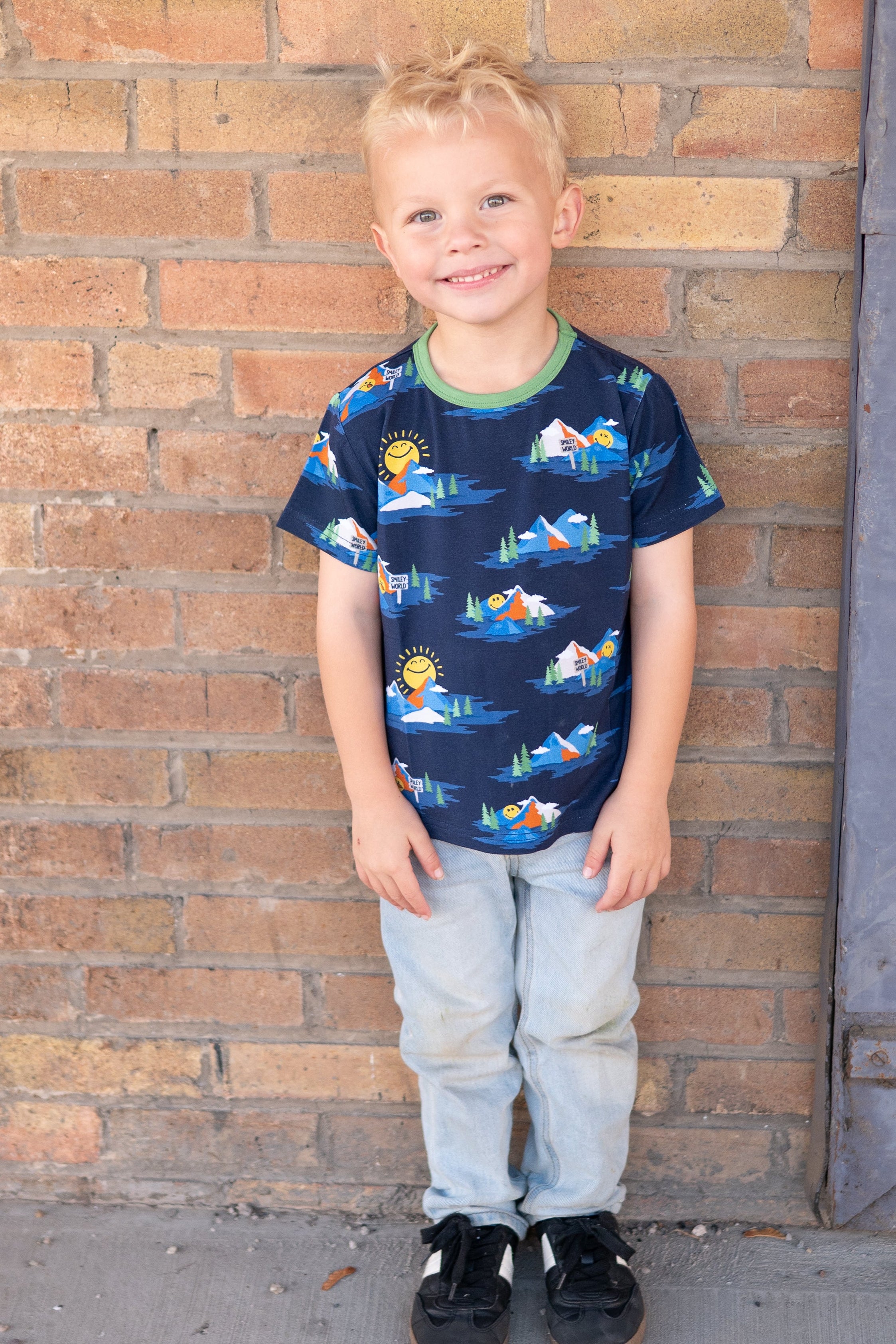 A young boy wears a navy blue t-shirt with a SmileyWorld mountain print and light wash jeans.