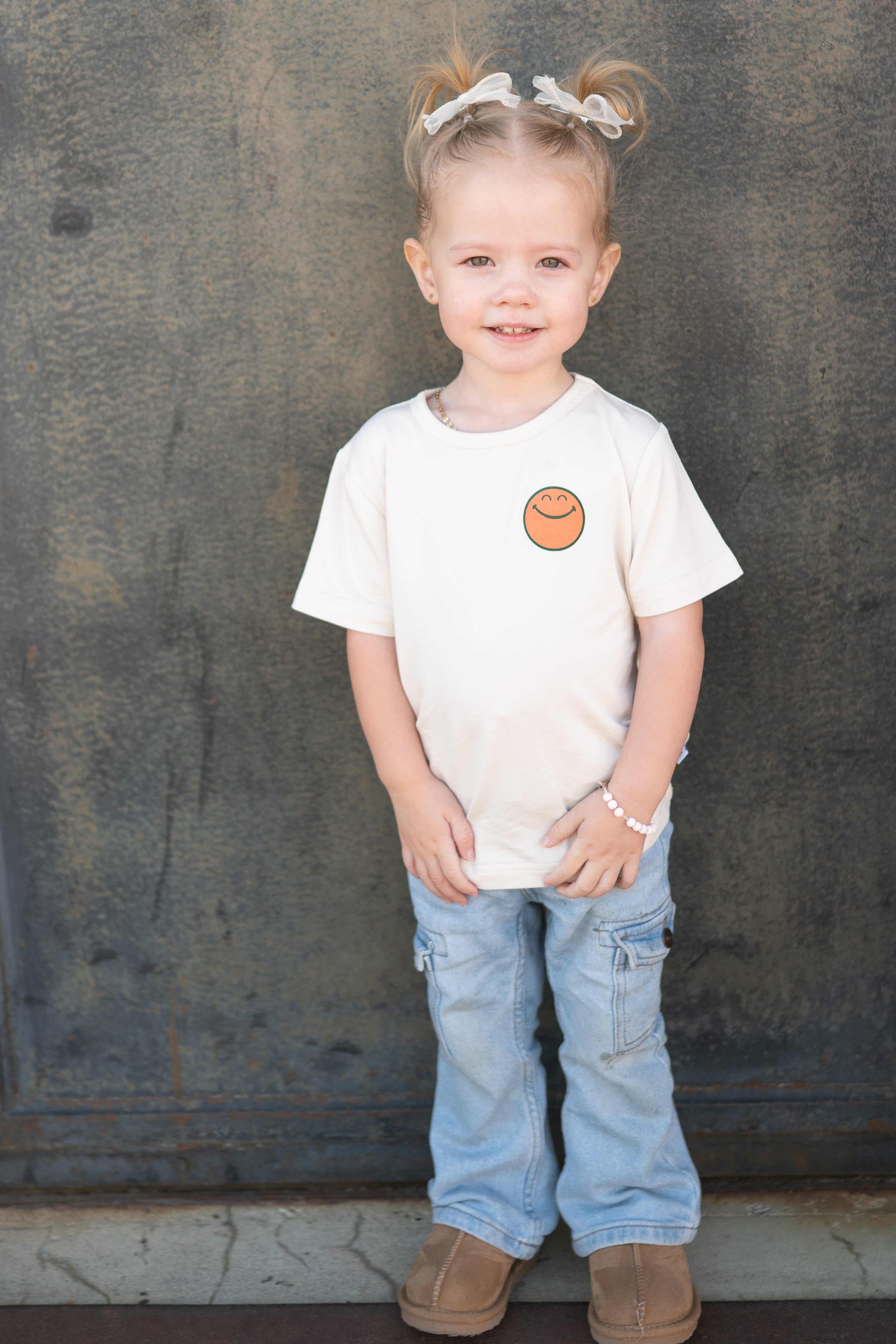 A young girl wears a cream t-shirt with a smiley face graphic.