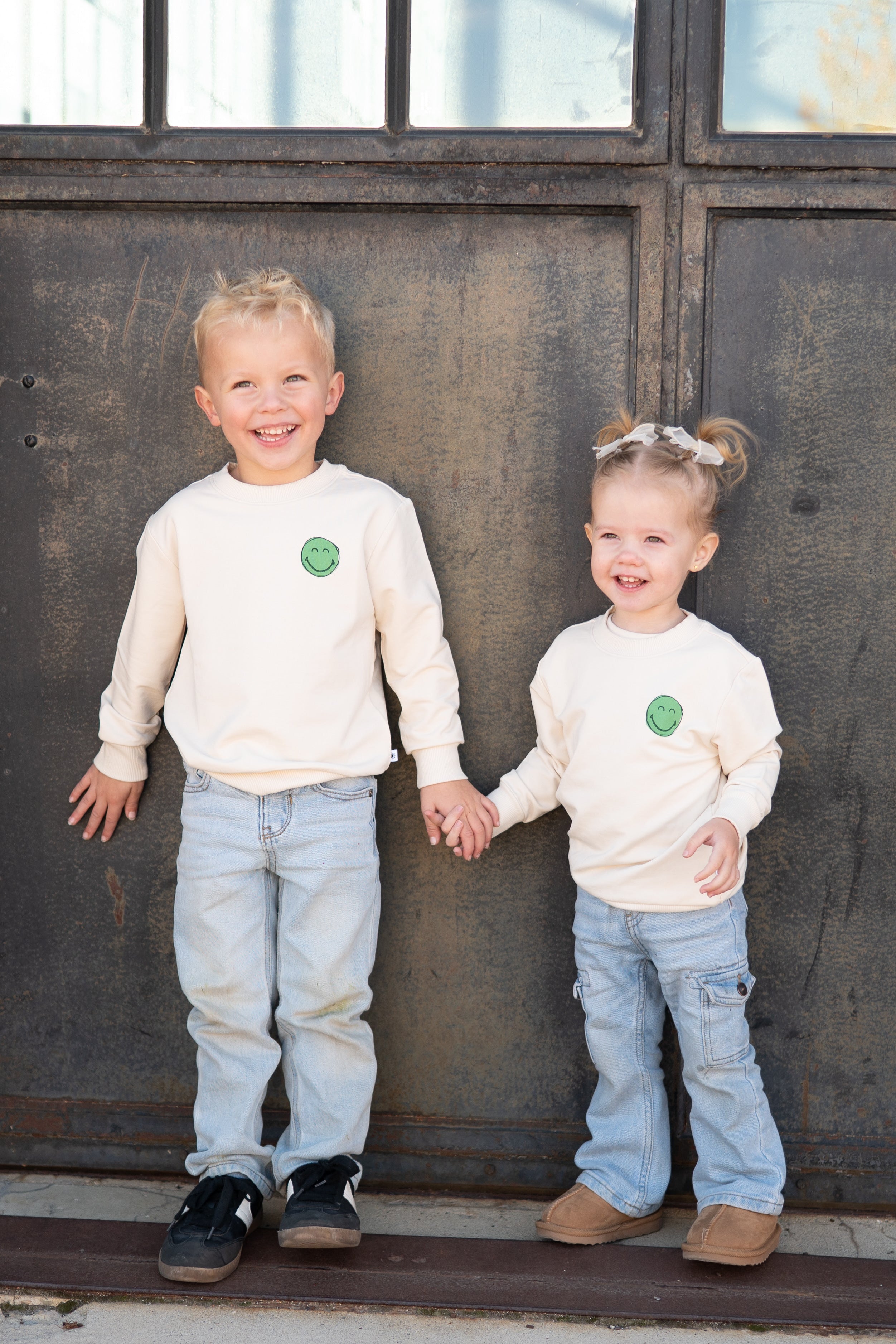 Two children in matching cream smiley face sweatshirts and light wash jeans.