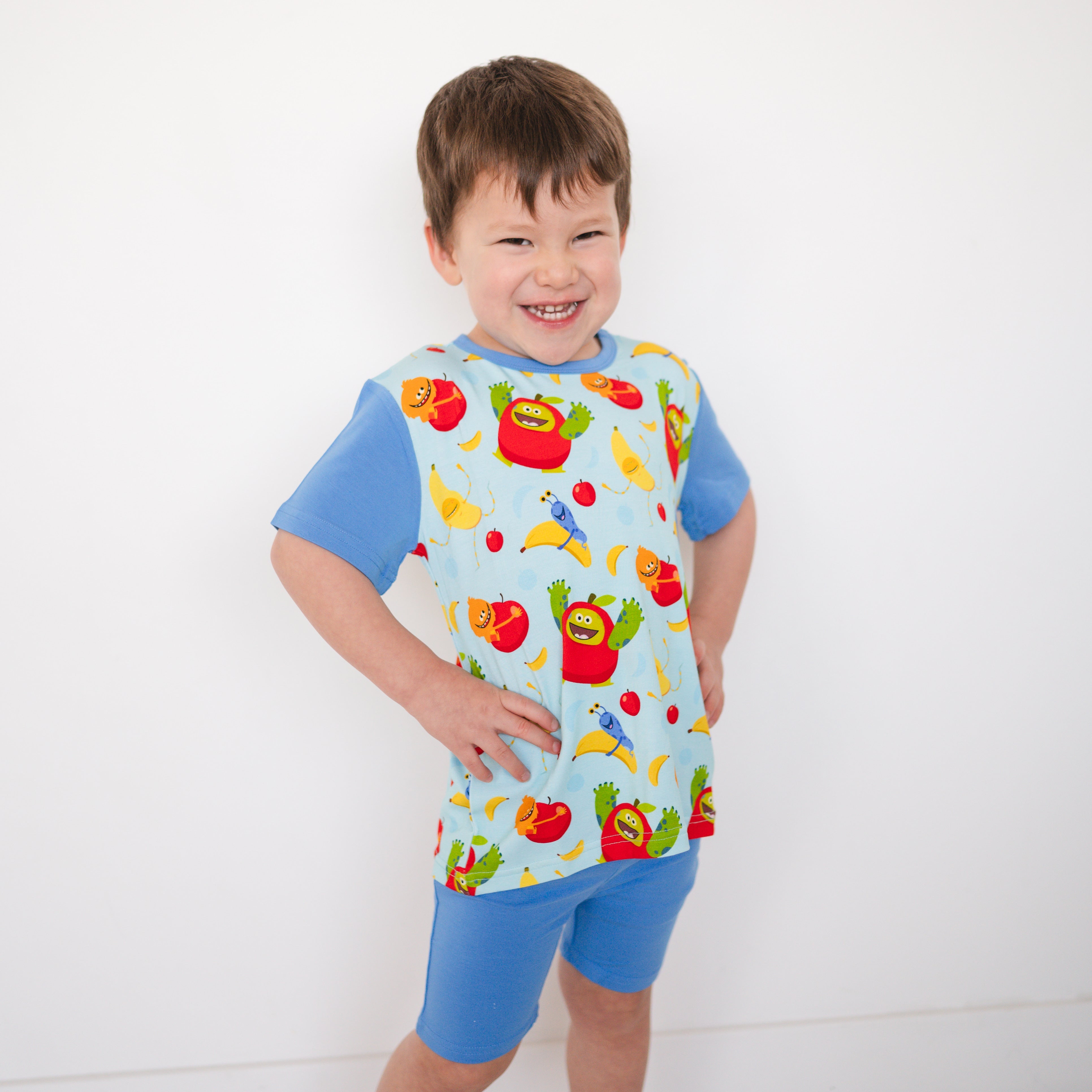 A young boy wears a light blue t-shirt with a colorful Apples & Bananas print and matching blue shorts.
