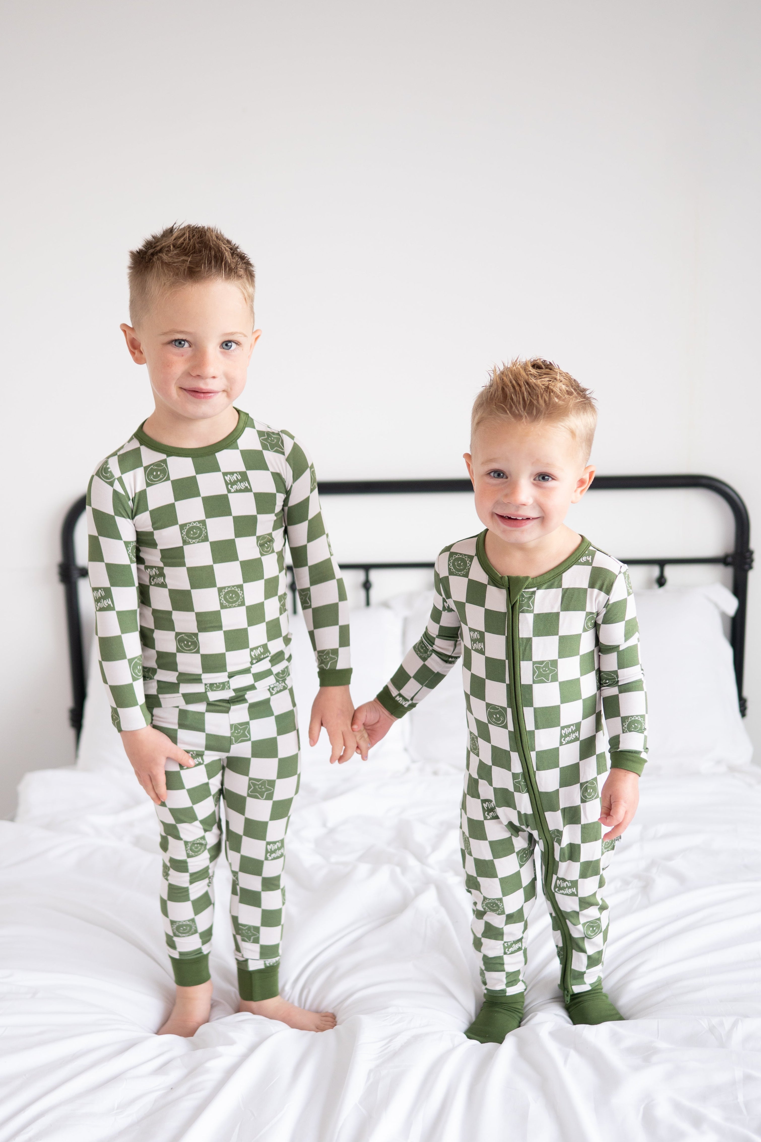 Two young boys in green and white checkered pajamas with smiley faces stand holding hands on a bed.