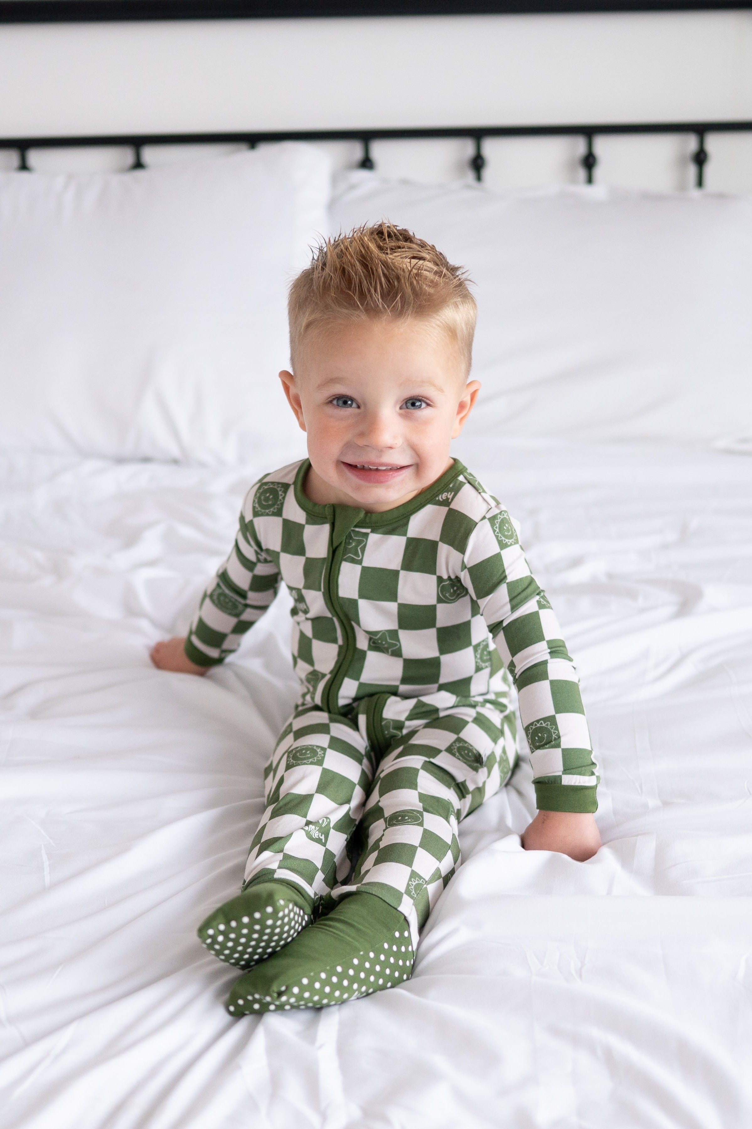 A baby boy smiles on a white bed wearing a green and white checkered bamboo pajama set with smiley faces.