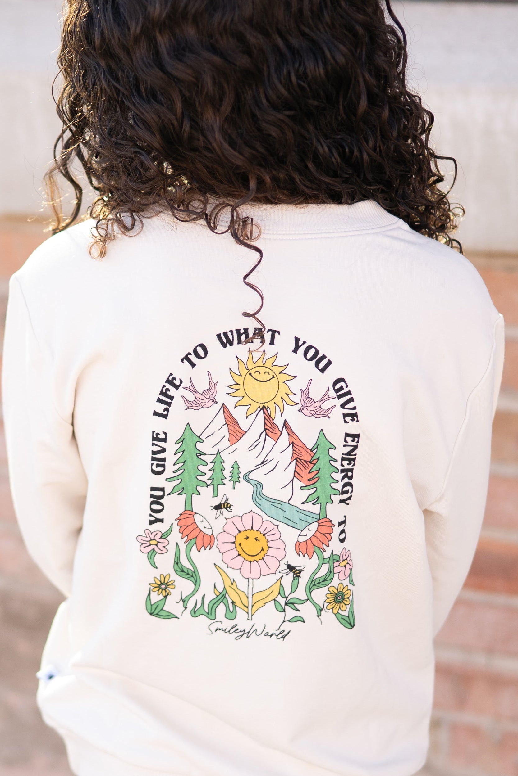 Cream crewneck sweatshirt with a colorful nature graphic and "You Give Life To What You Give Energy To" text.
