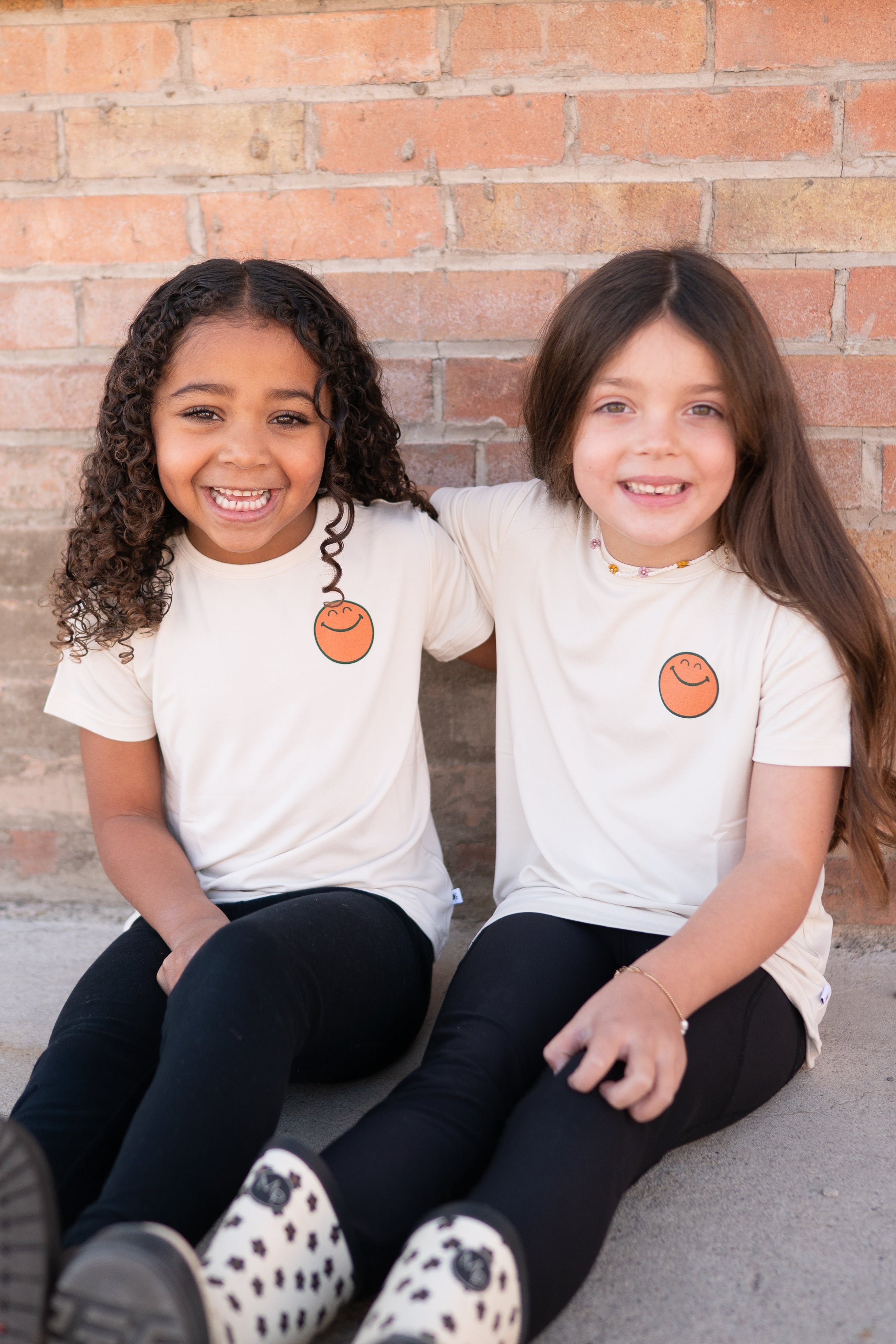 Two young girls in cream t-shirts with smiley face graphics sit in front of a brick wall.