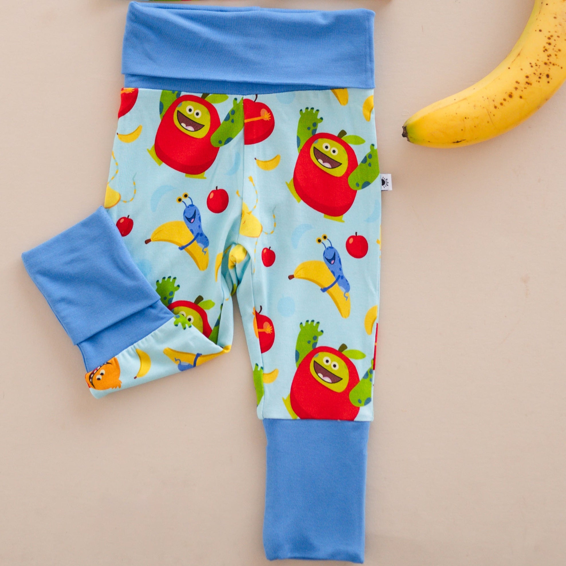 Baby pants with a light blue background featuring a pattern of cartoon apples, bananas, and monsters.