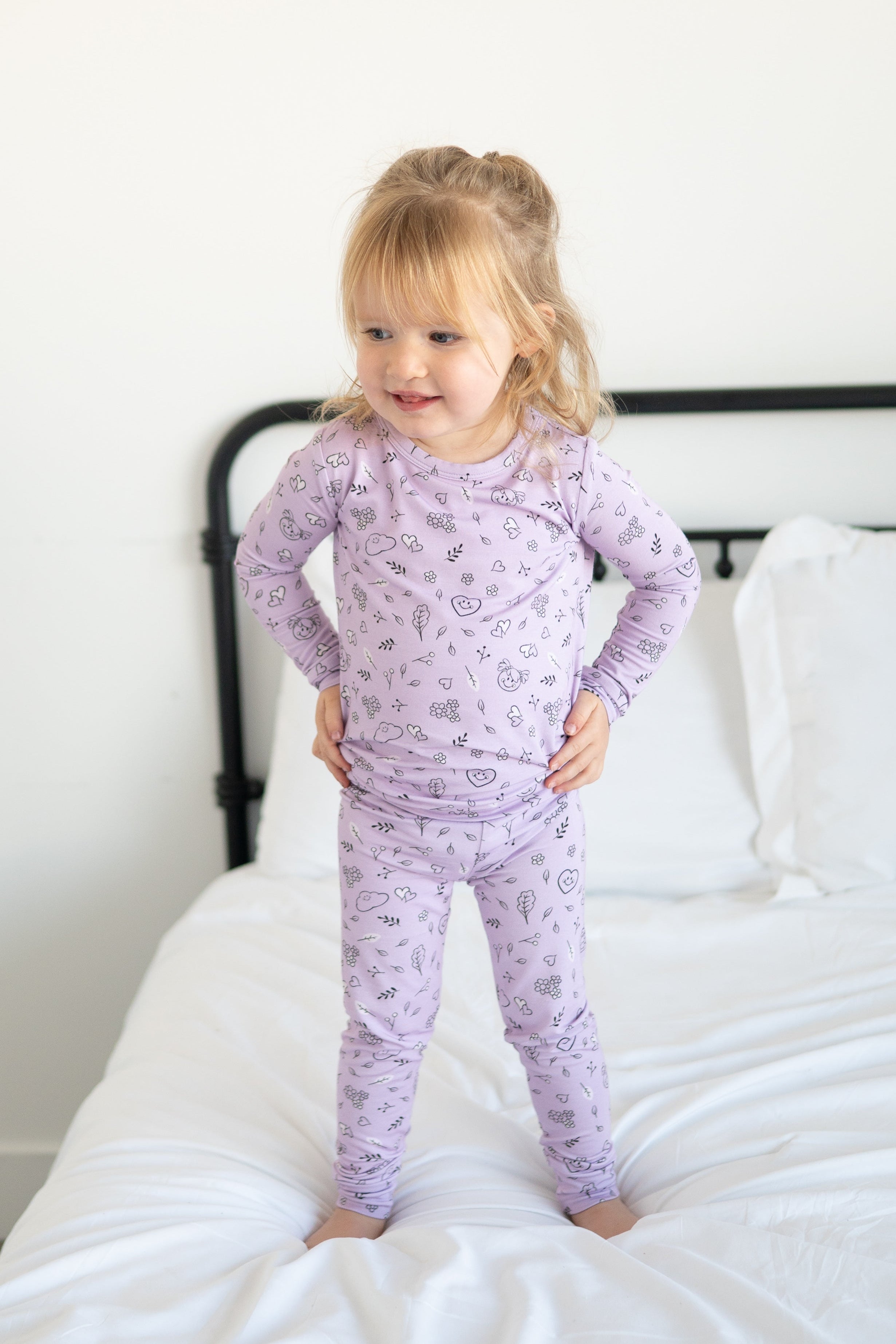 A young girl stands on a bed wearing lilac pajamas with a doodle print.