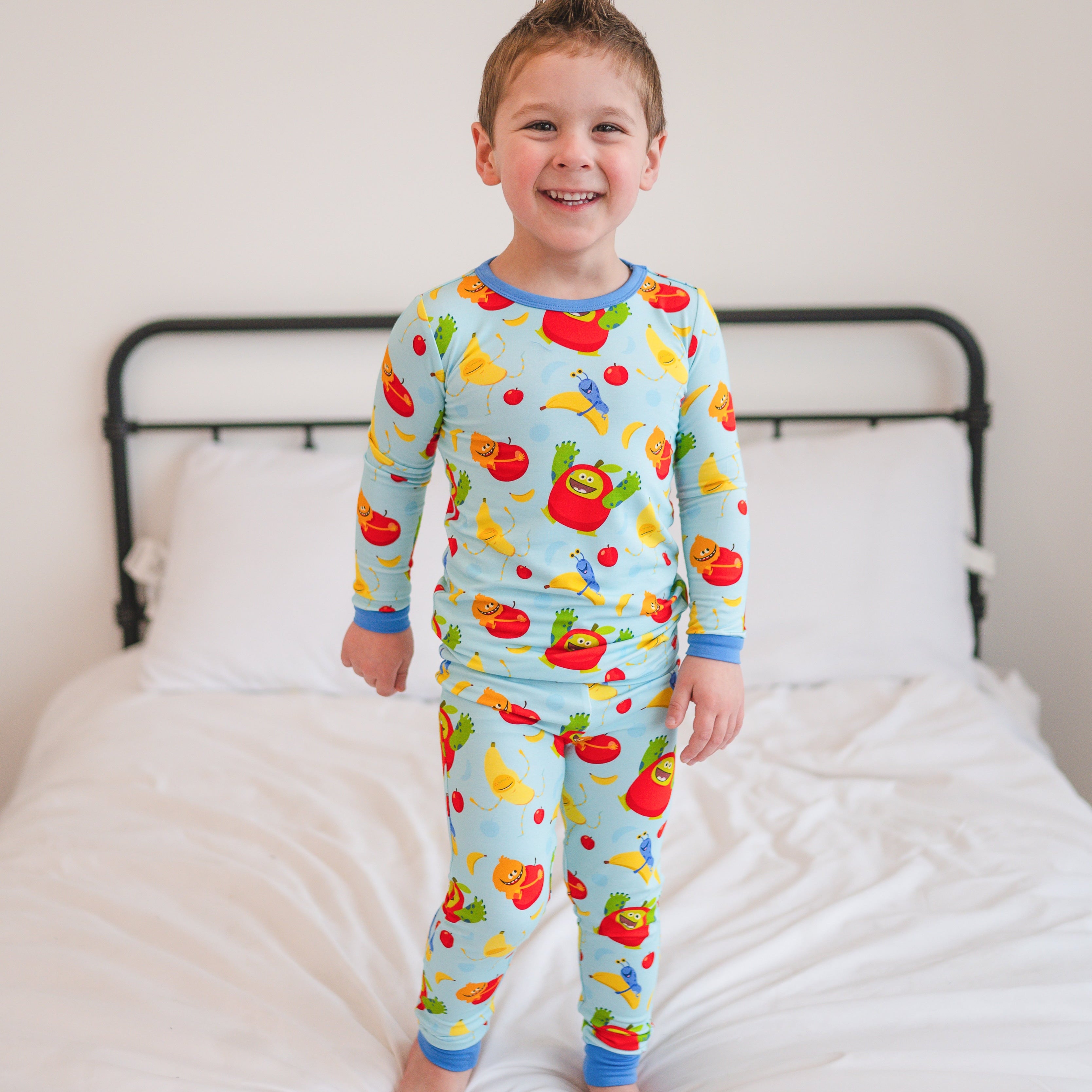 A young boy smiles wearing light blue pajamas with a print of cartoon apples and bananas.