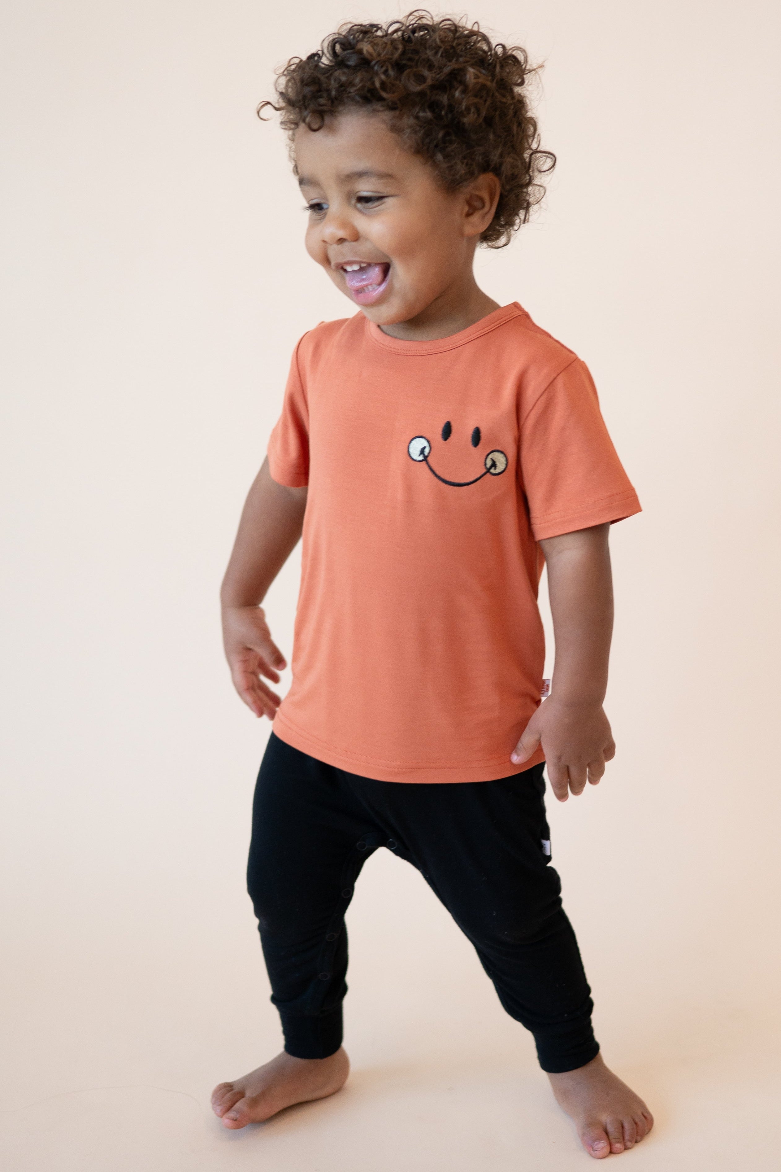 A young child wears a rust-colored t-shirt with an embroidered smiley face and black pants.