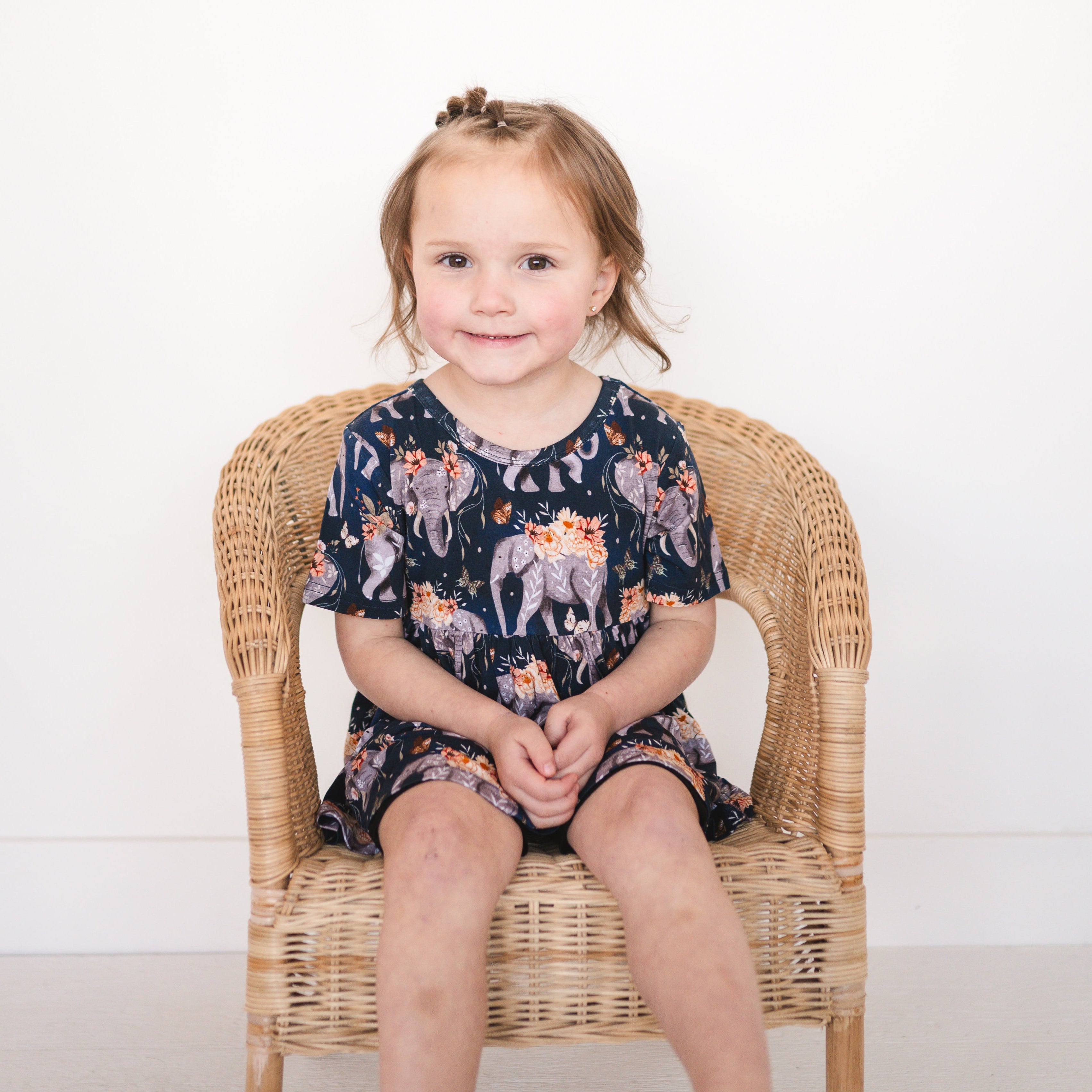 A young girl sits in a wicker chair wearing a navy peplum top with an elephant and floral print.