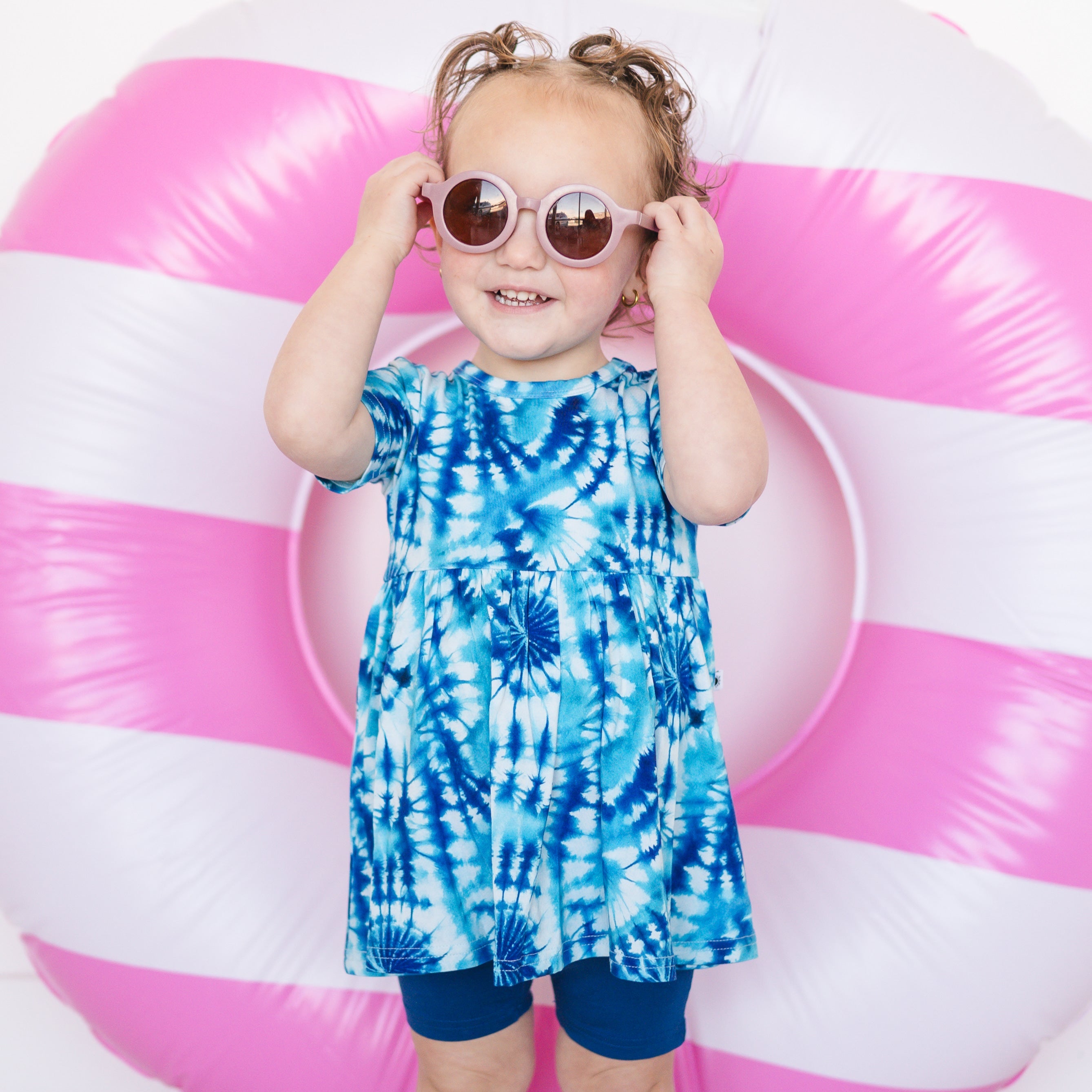 Toddler girl in a blue tie-dye peplum top and navy shorts, wearing pink sunglasses.