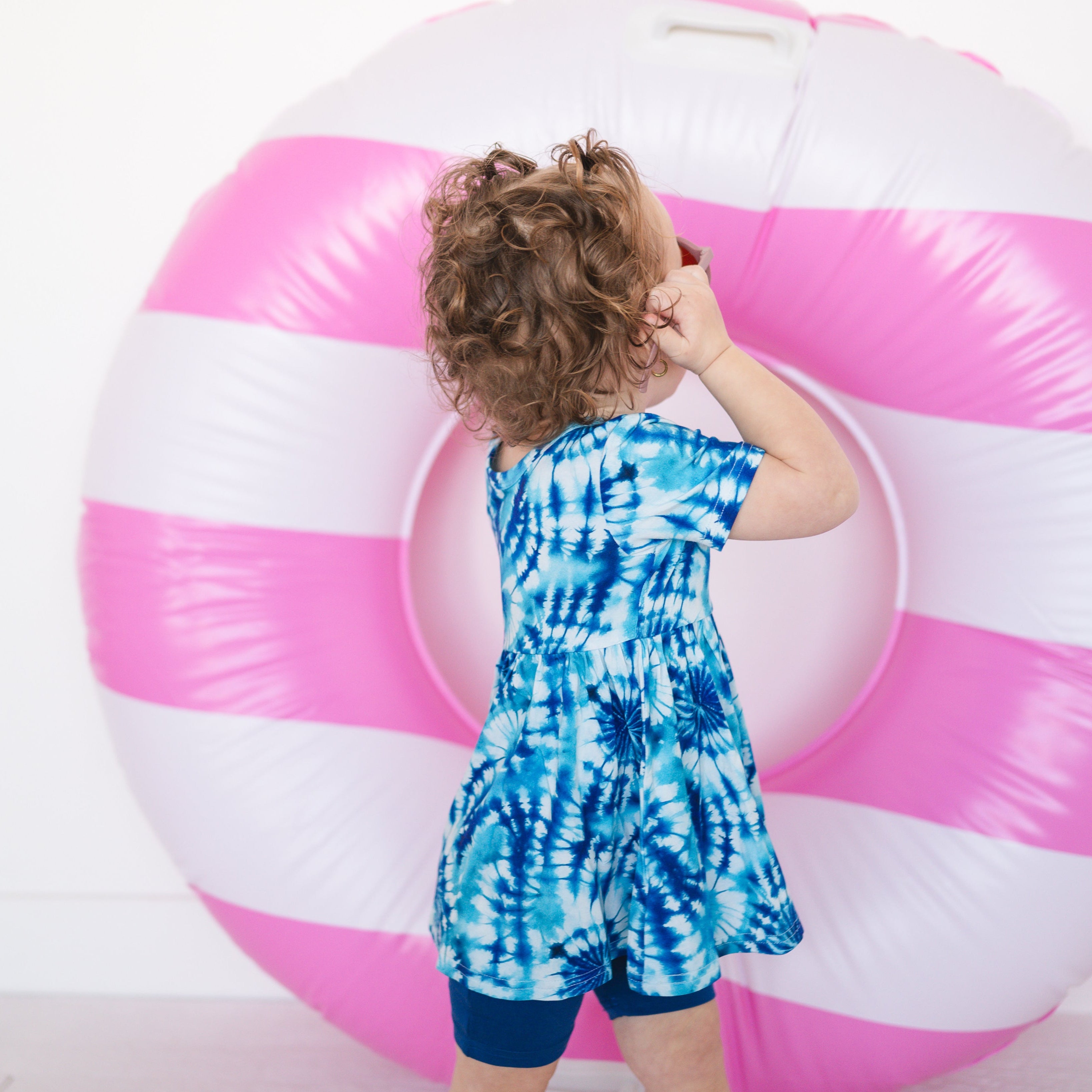 A young child wears a blue tie-dye peplum top and blue shorts in front of a pink and white inflatable ring.