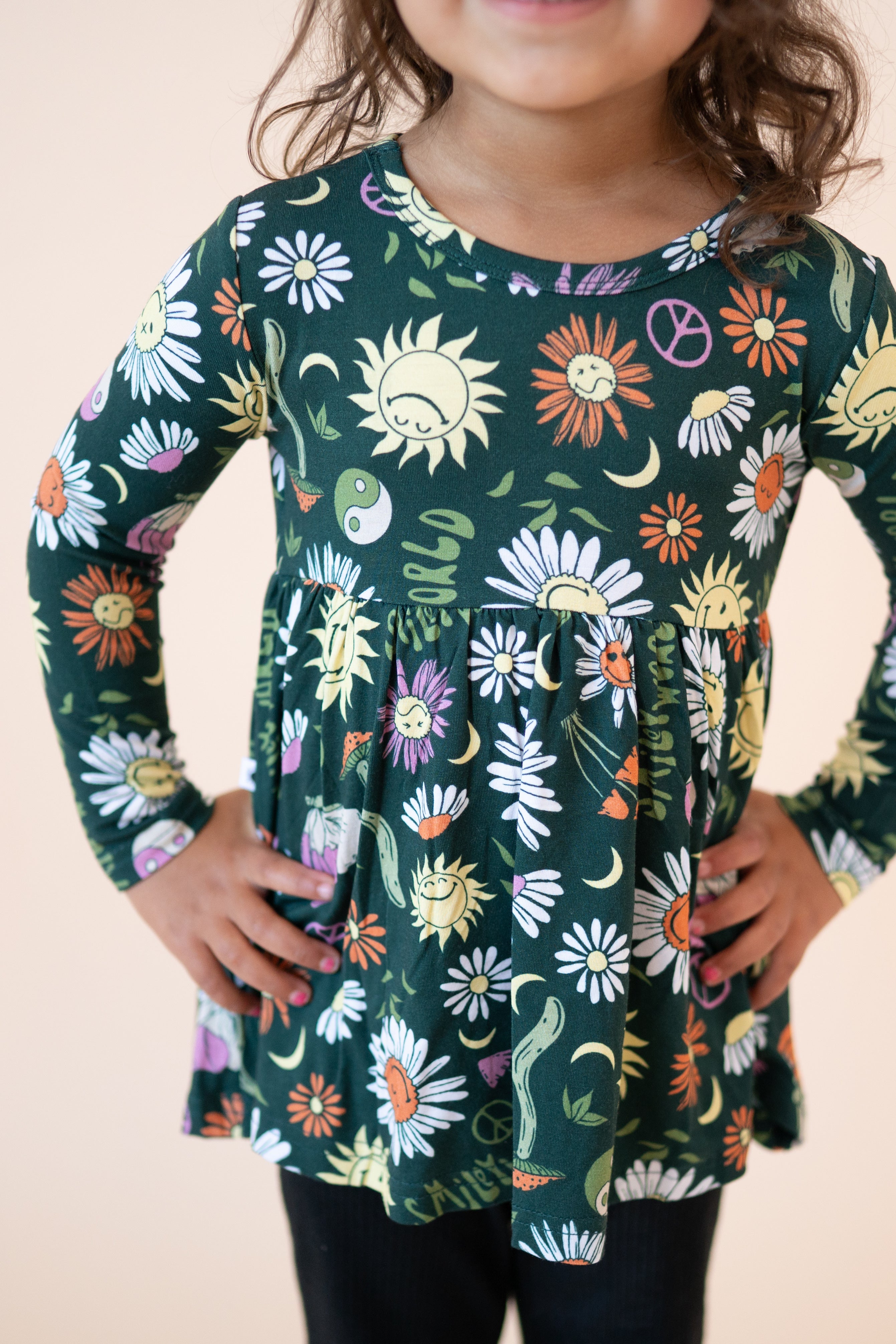 A girl wears a long-sleeve green peplum top with a groovy floral and smiley sun print.