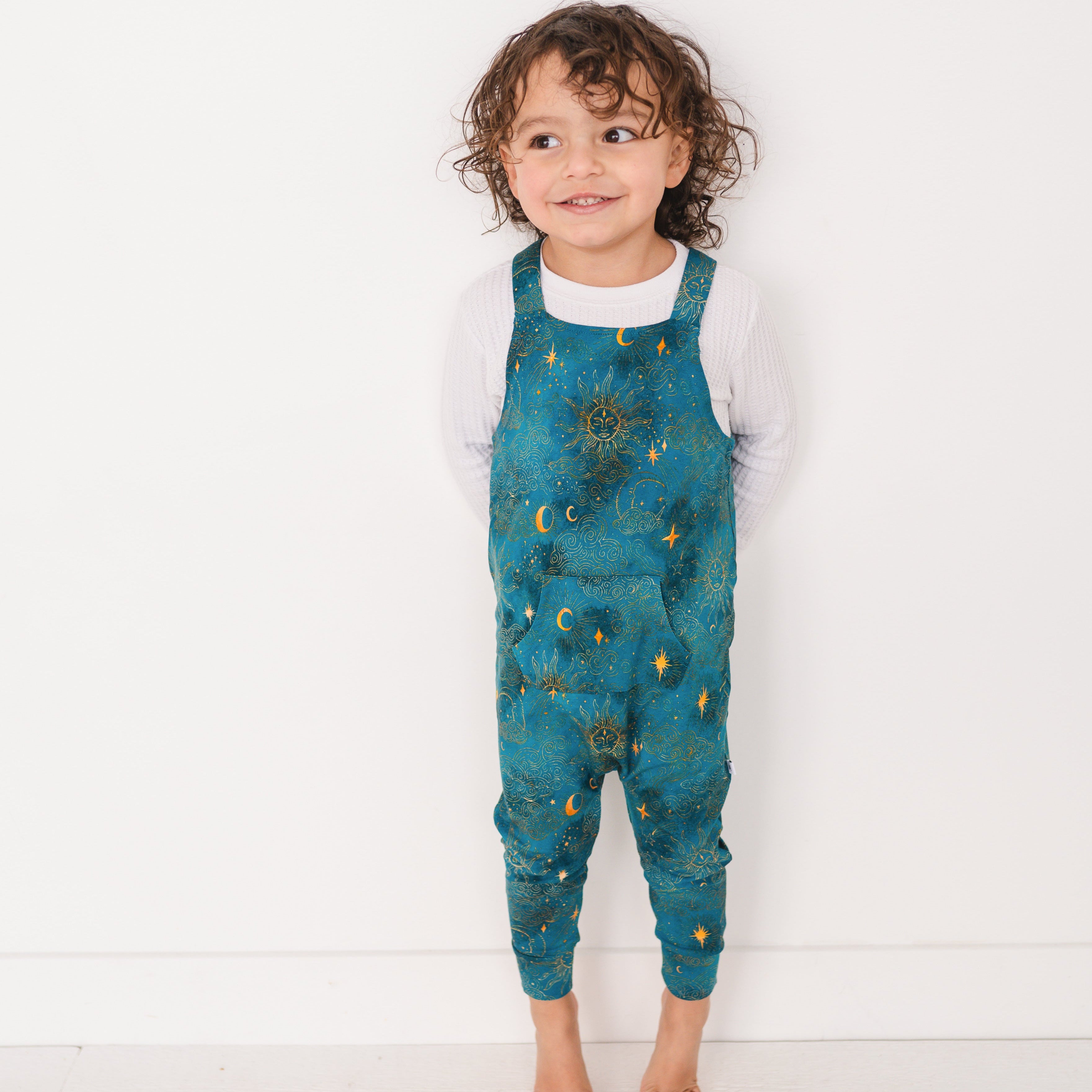 A young child wears teal overalls with a celestial sun and moon print.