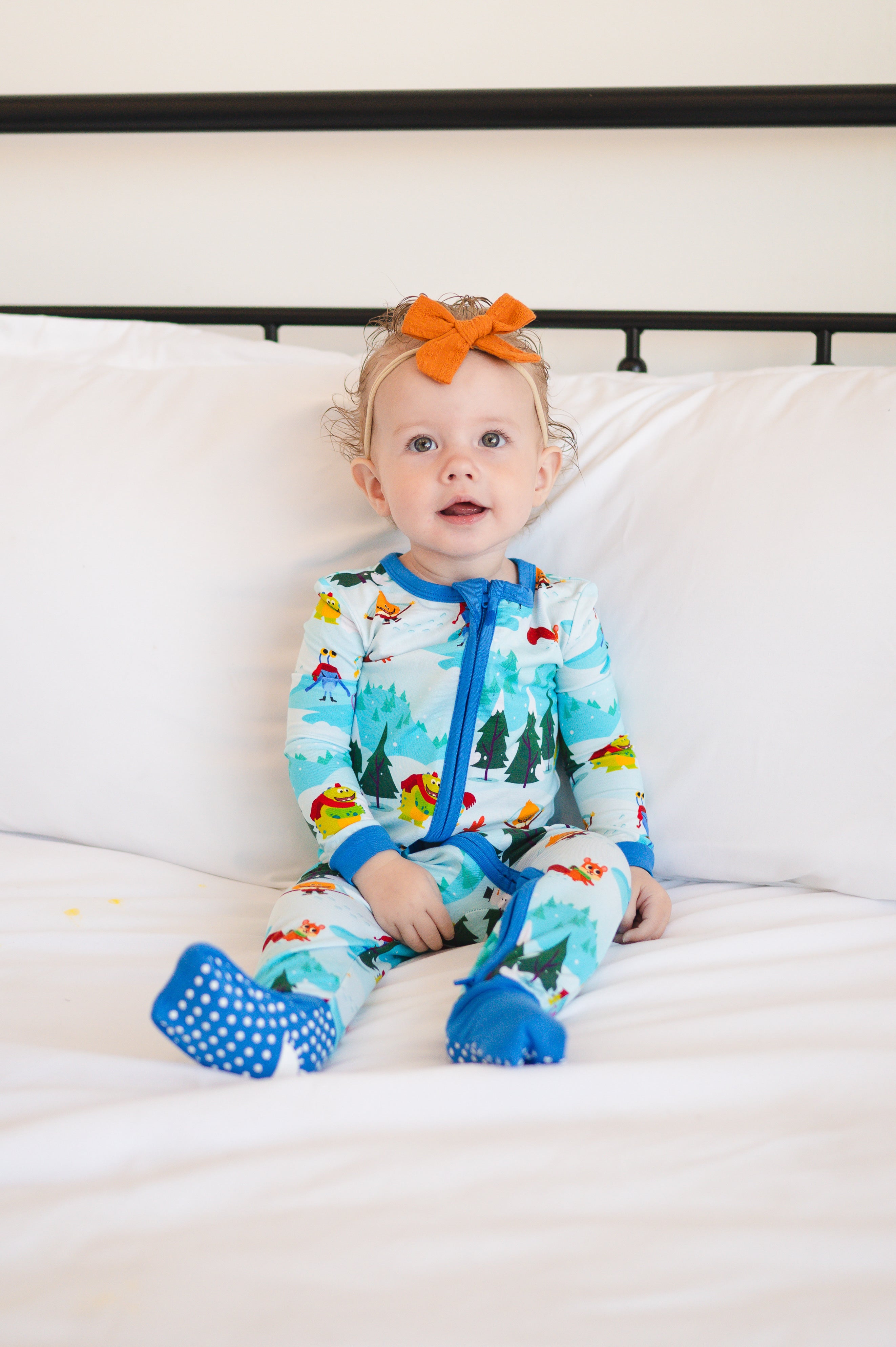 A baby girl wearing a blue snow-themed pajama set with an orange bow sits on a white bed.