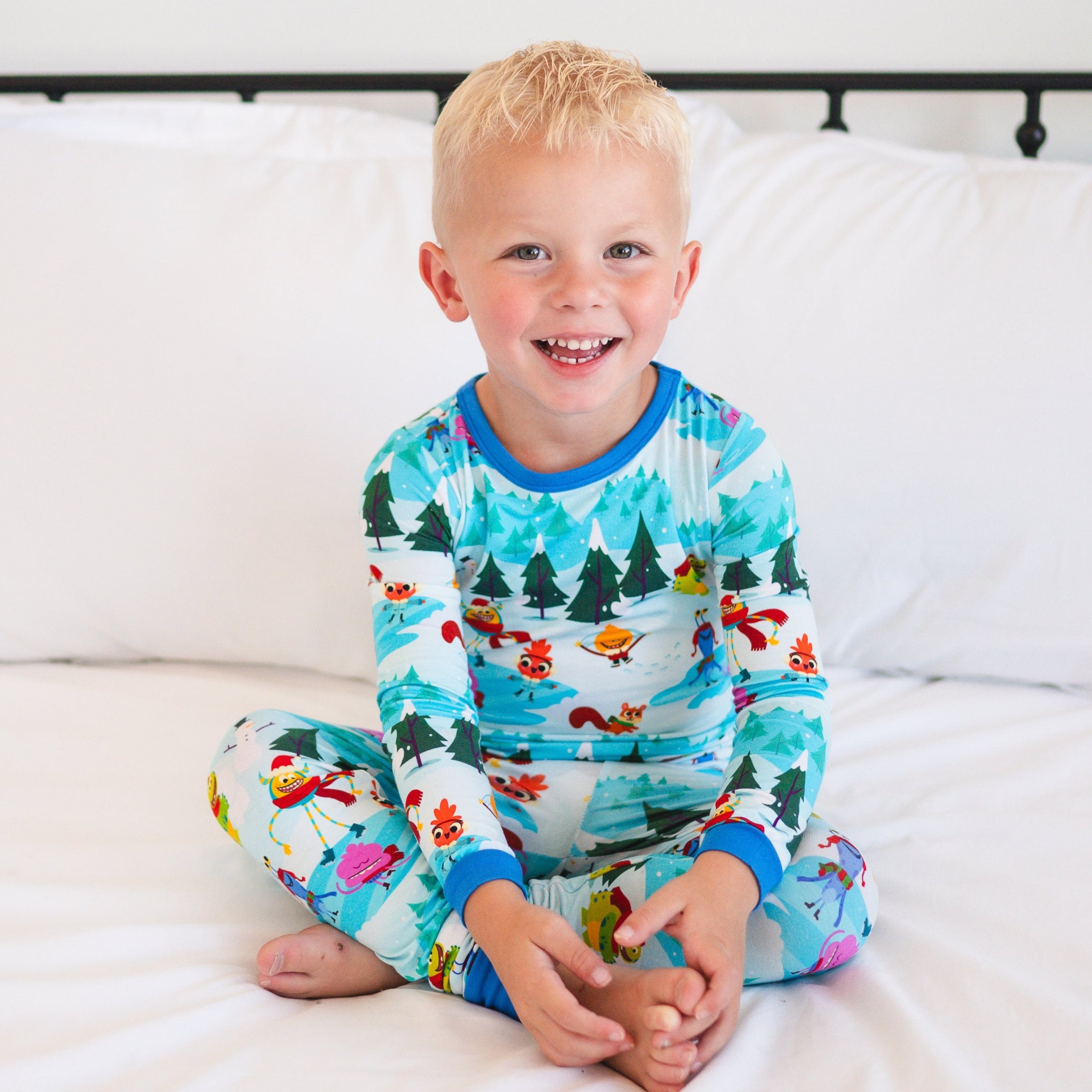 A young boy in blue pajamas with a winter forest and cartoon character print sits cross-legged on a white bed.