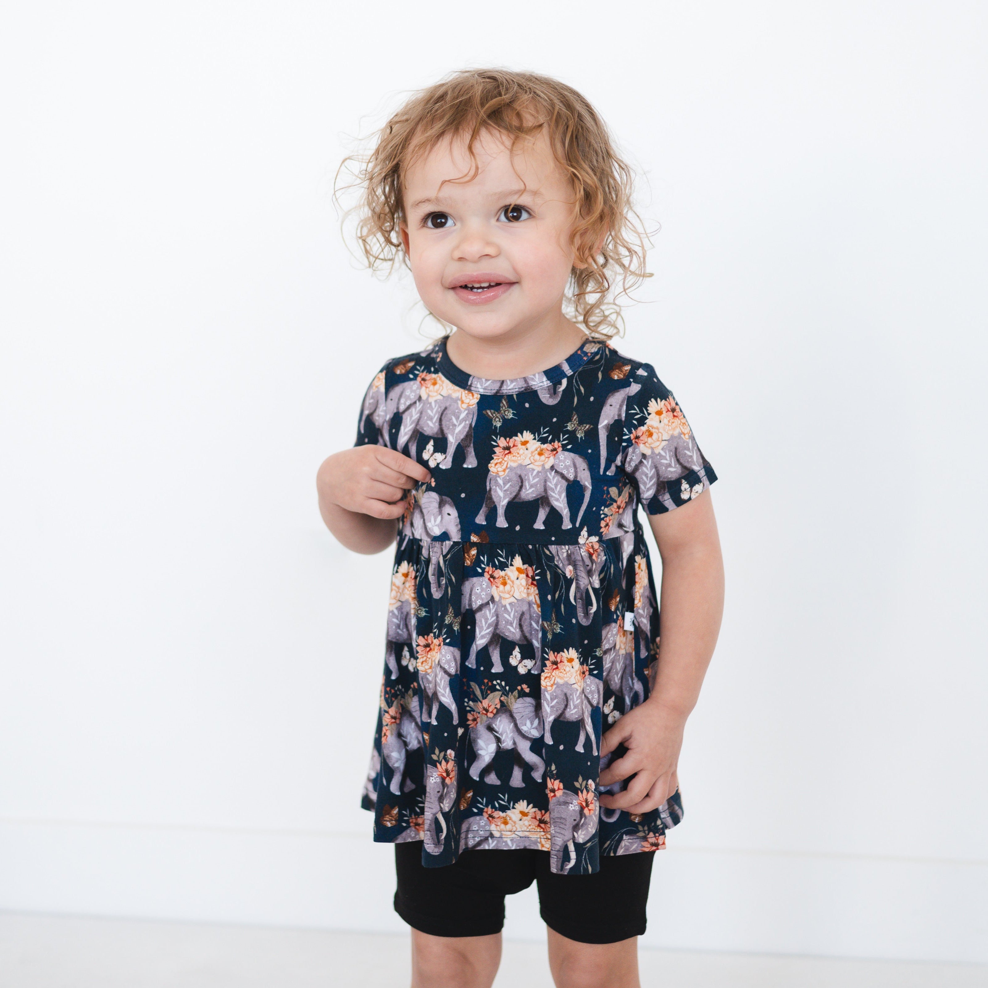 A young girl wears a navy peplum top with an elephant and floral print and black shorts.