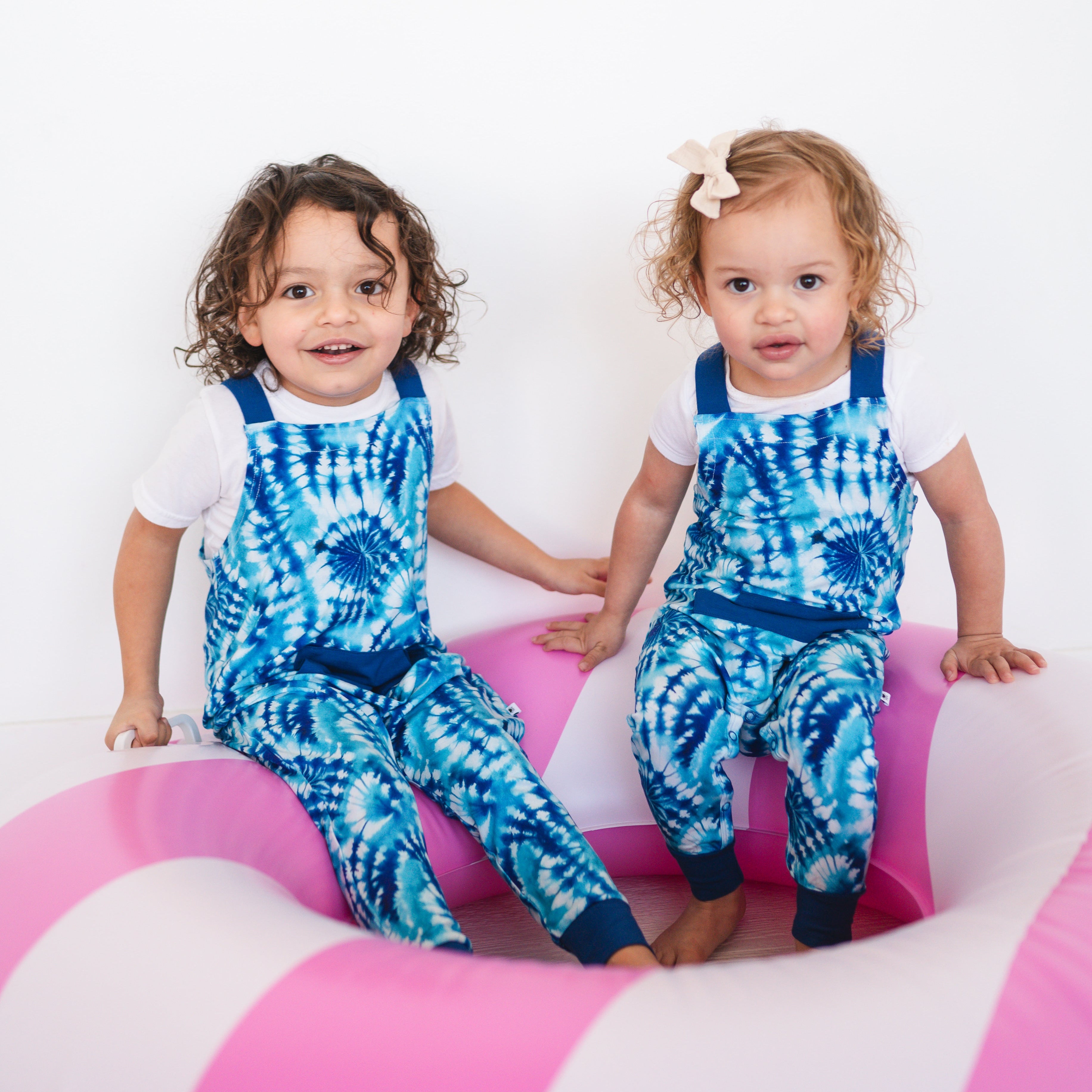 Two children in blue tie-dye overalls sit in a pink and white inflatable.