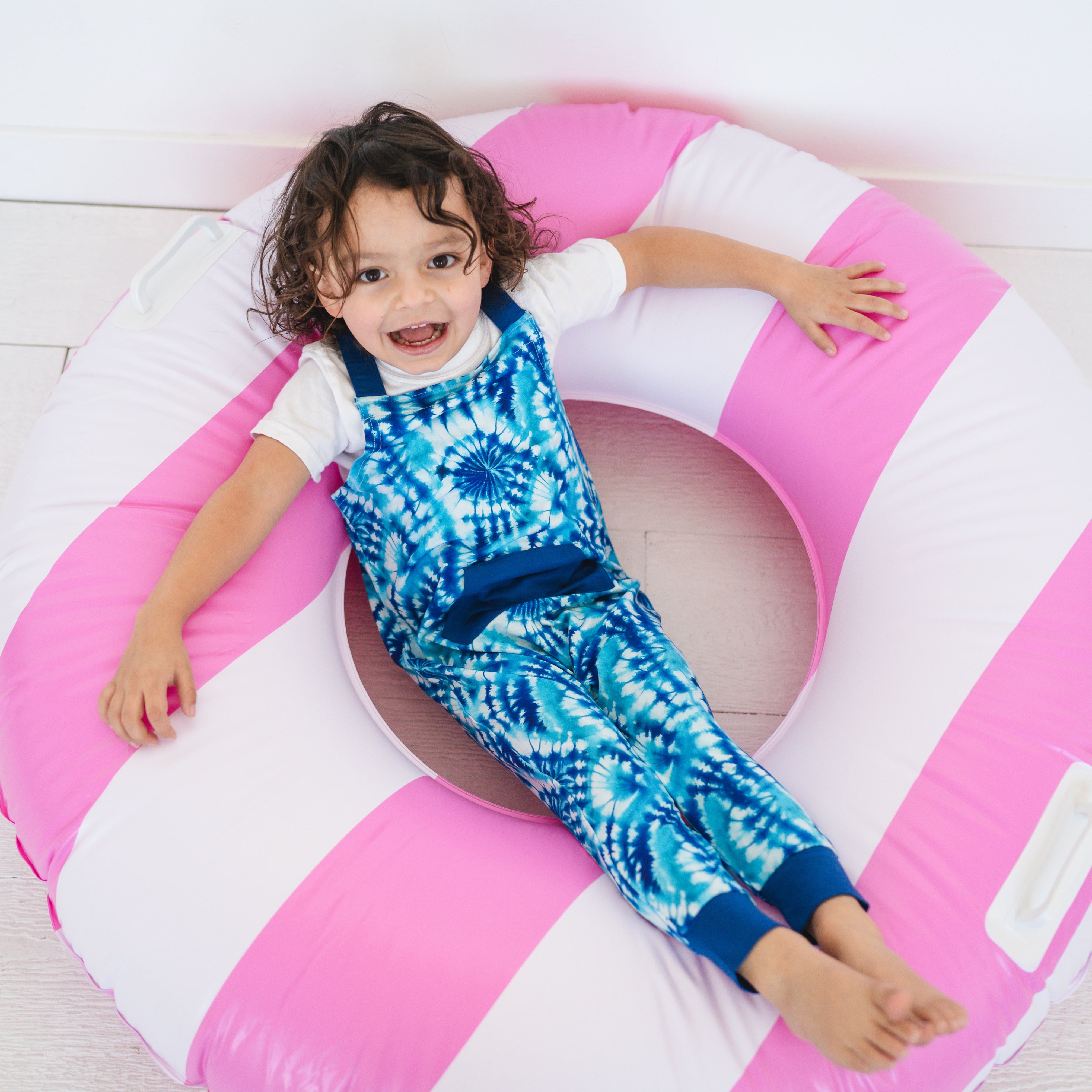 A child wears blue tie-dye overalls while sitting in a pink and white inflatable ring.