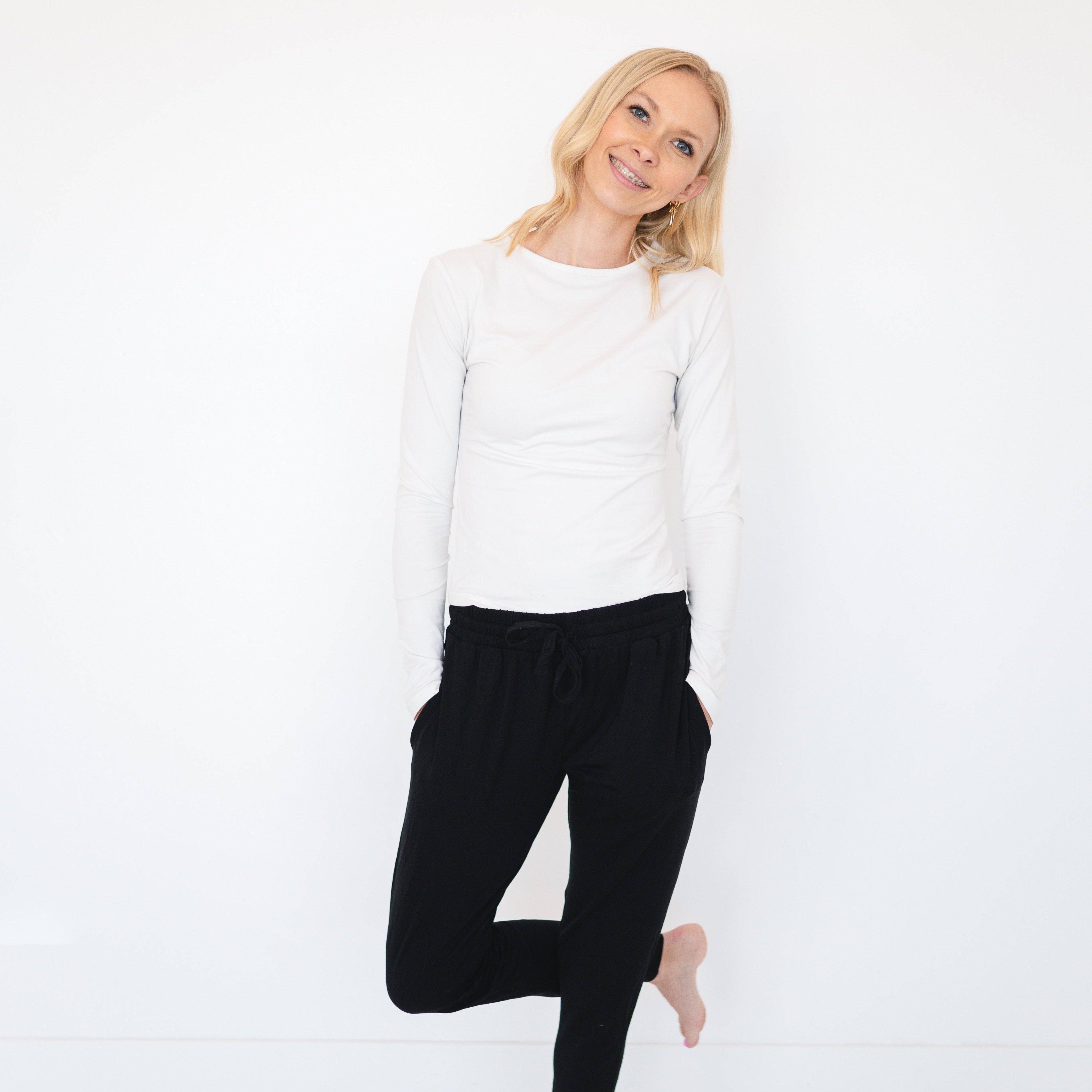 A woman in a white long-sleeve shirt and black joggers stands with one leg bent.