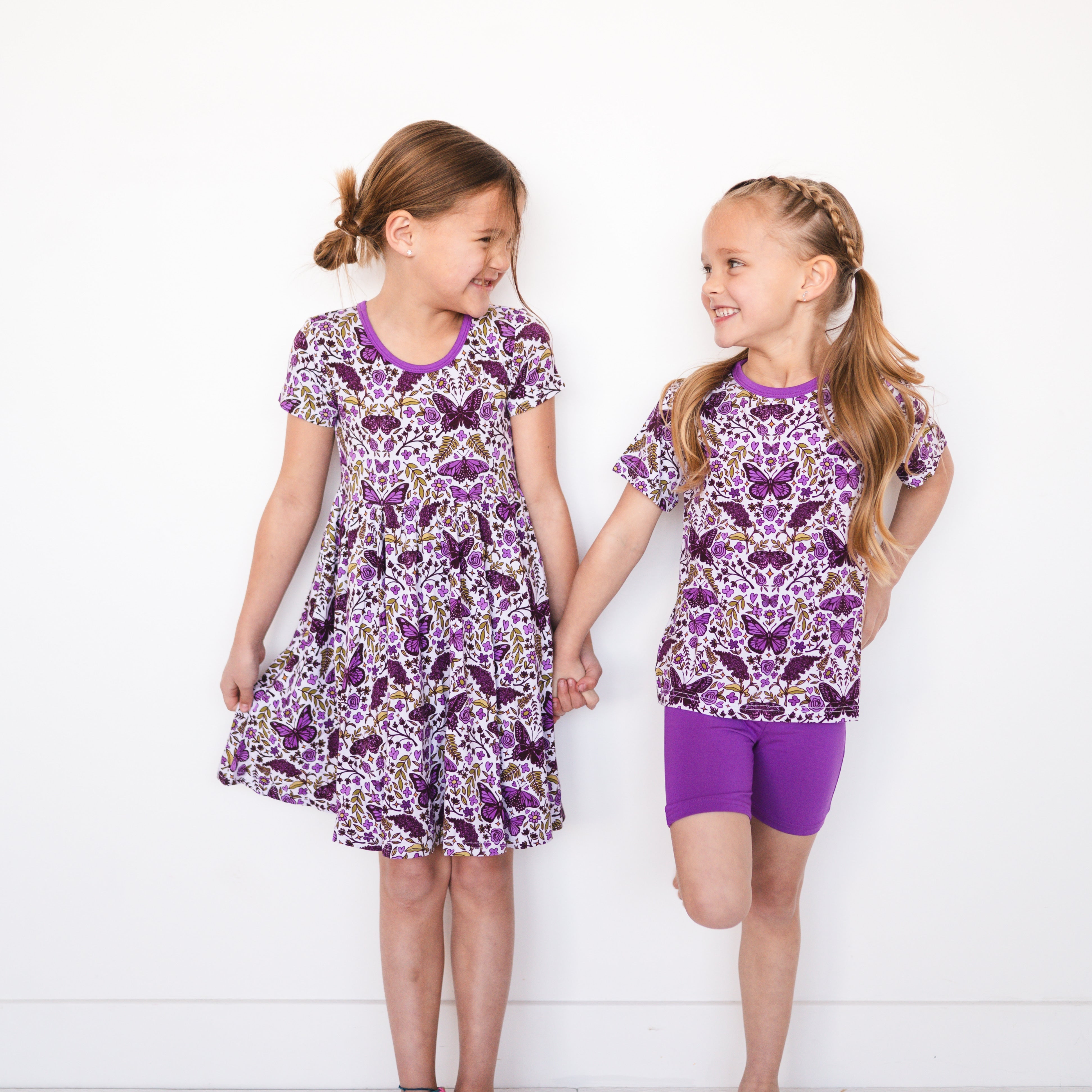 Two girls in matching purple butterfly print outfits stand holding hands.