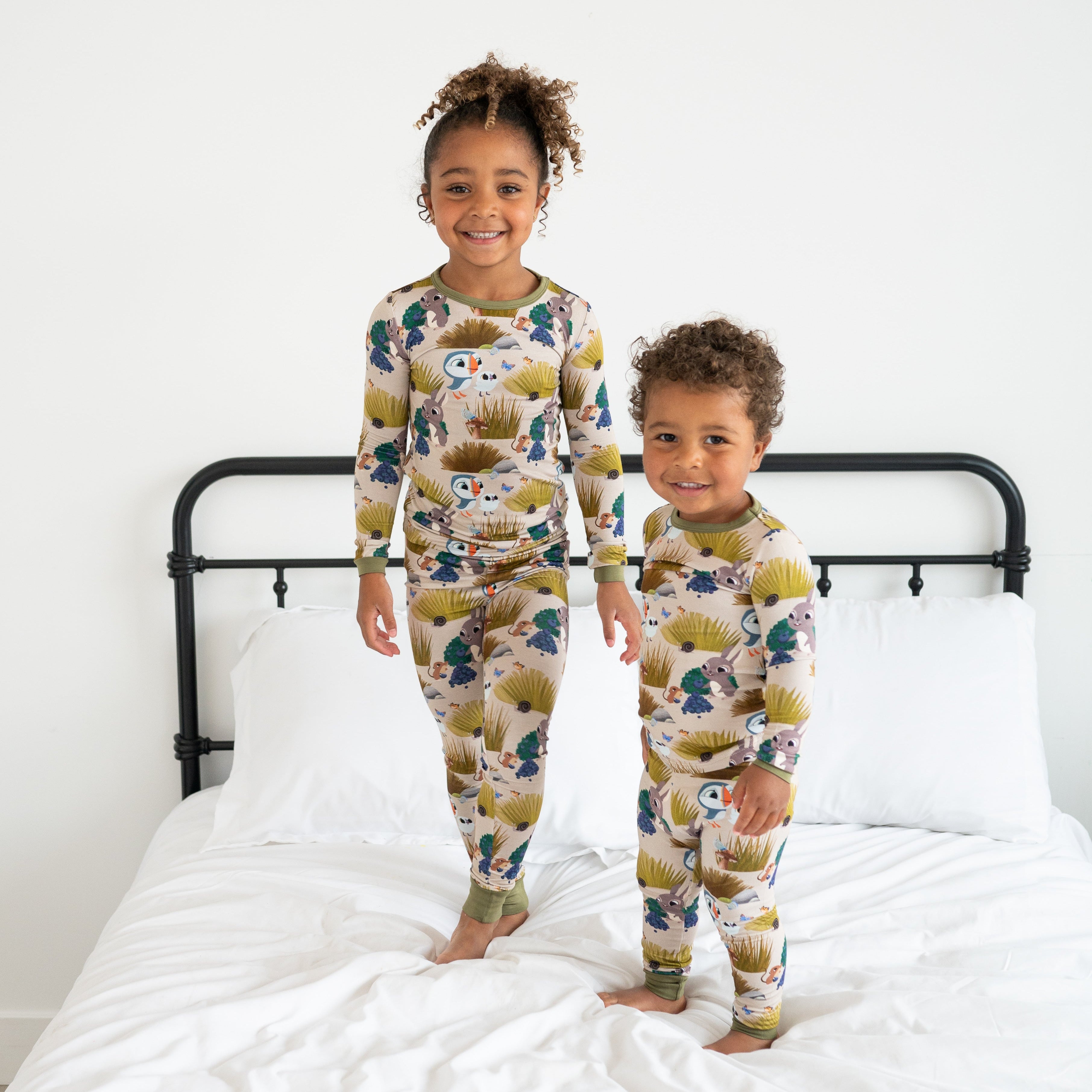 Two children in matching Puffin Rock™ bamboo pajamas stand on a bed.