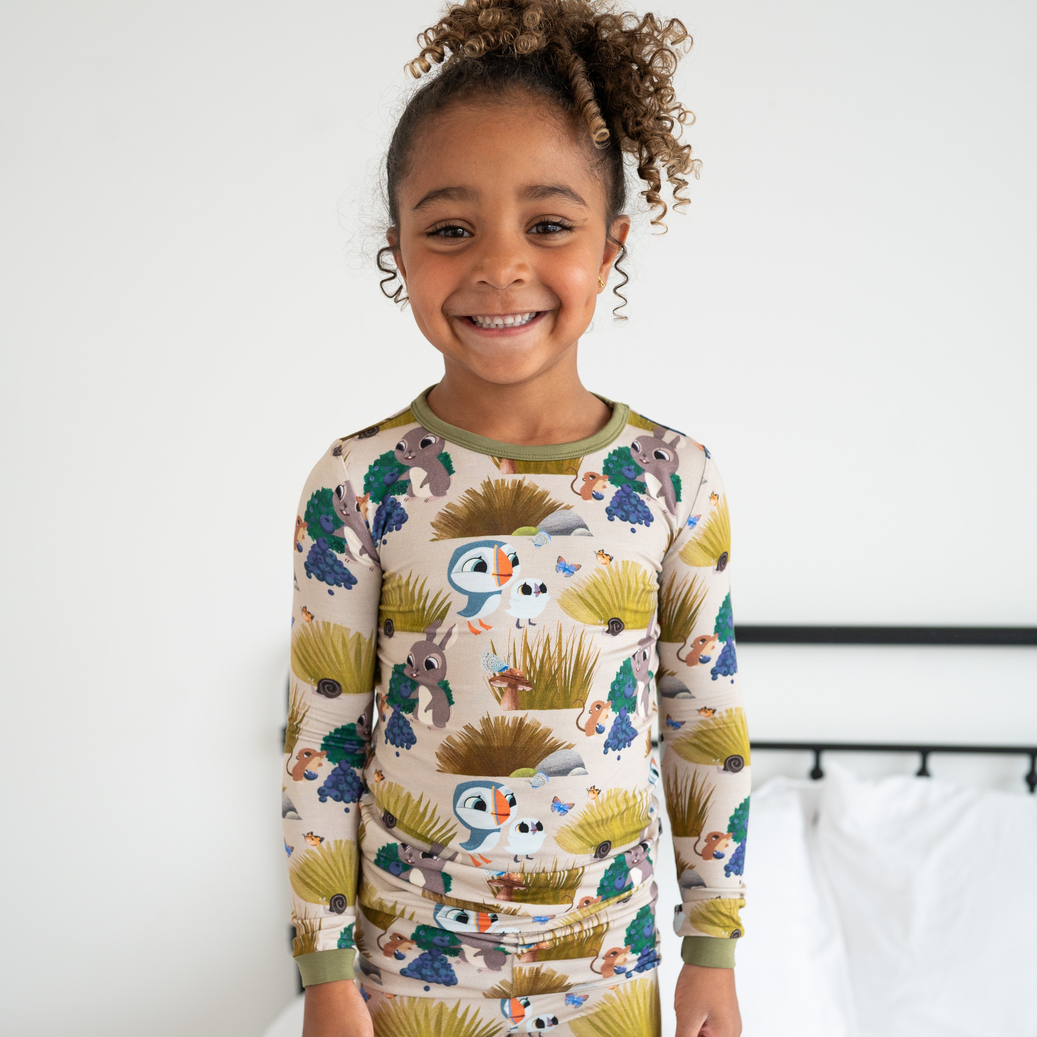 A young girl smiles wearing a long-sleeved pajama set with a puffin and woodland creature print.