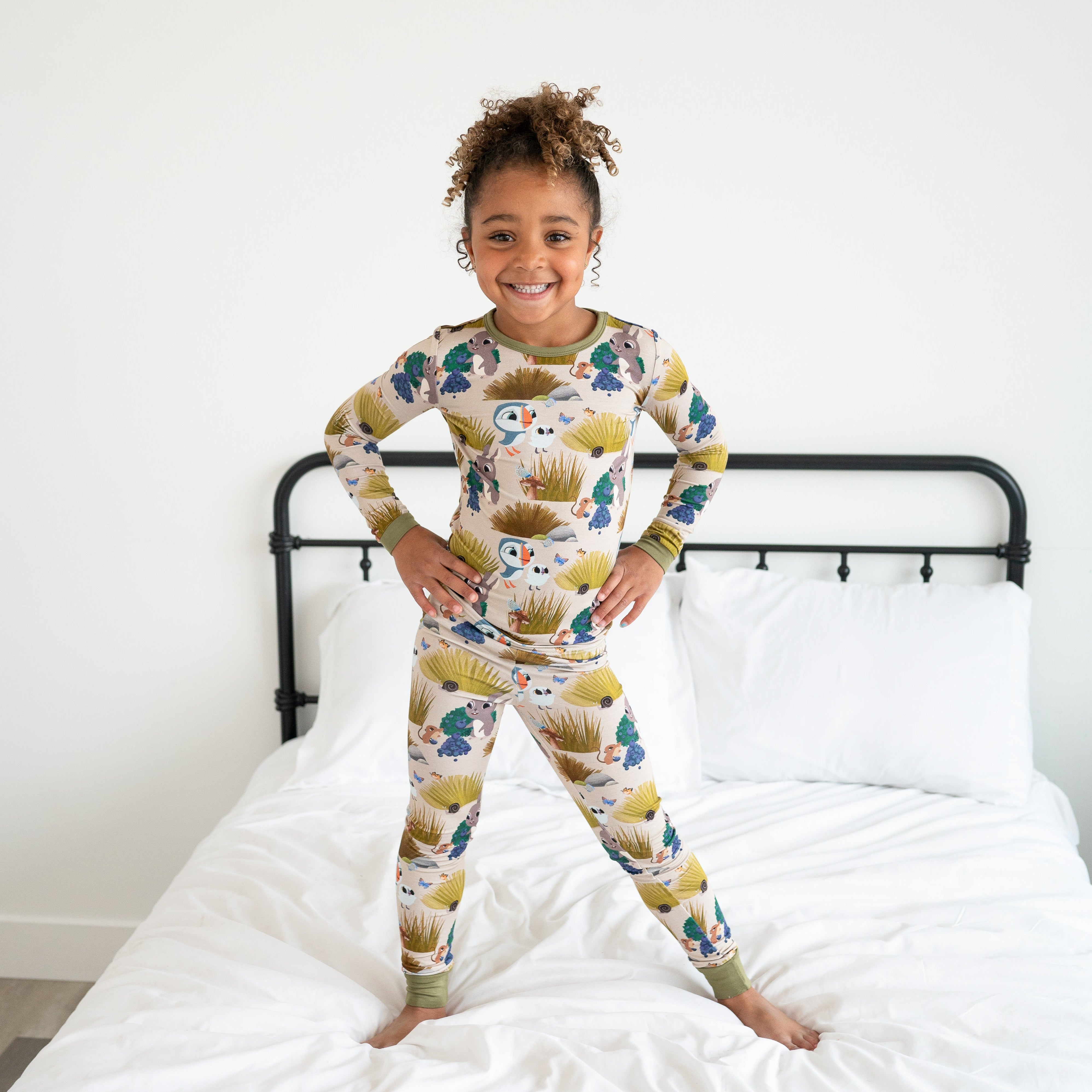 A young girl stands on a bed wearing a long-sleeved pajama set with a woodland animal print.