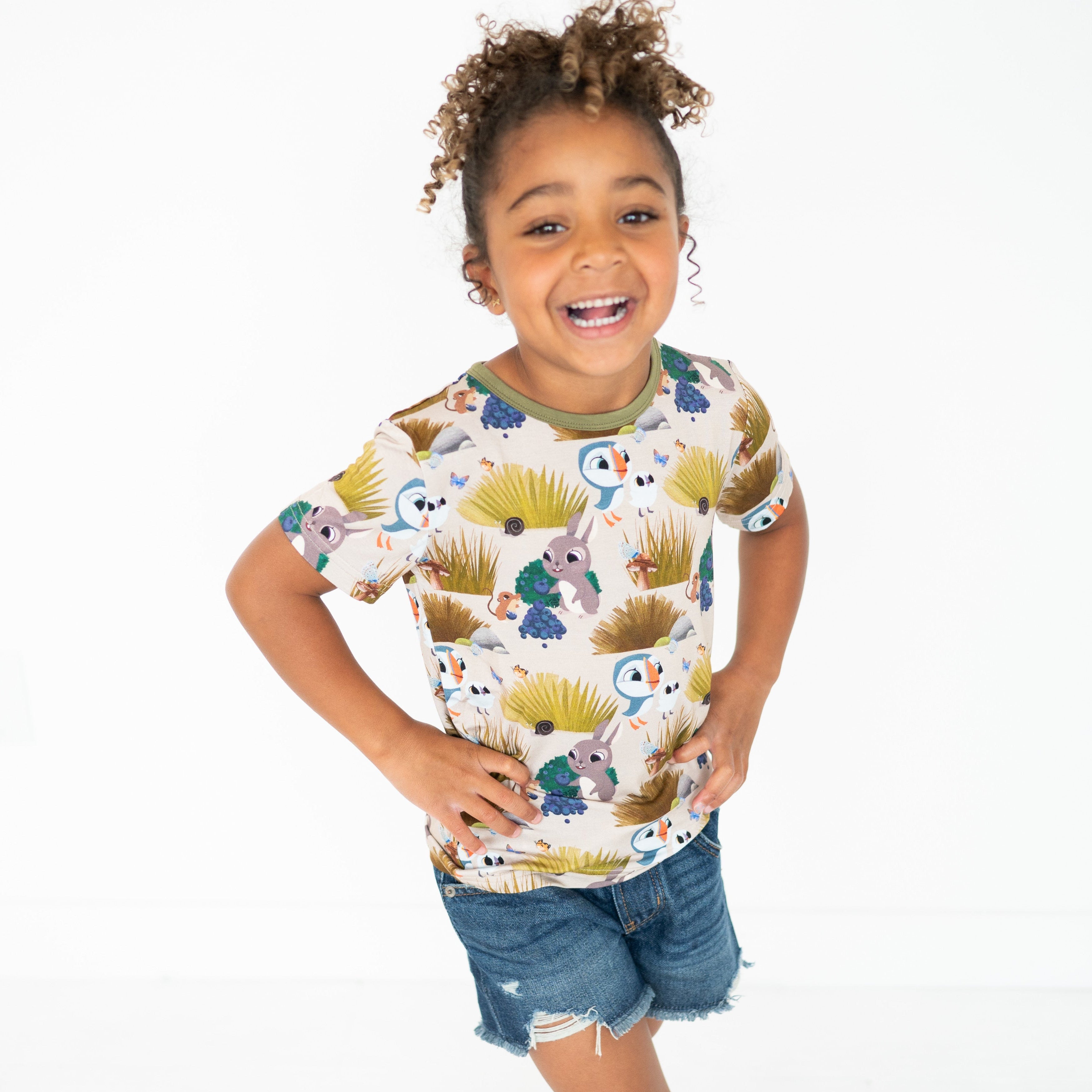 A young girl smiles wearing a tan t-shirt with a Puffin Rock print and denim shorts.