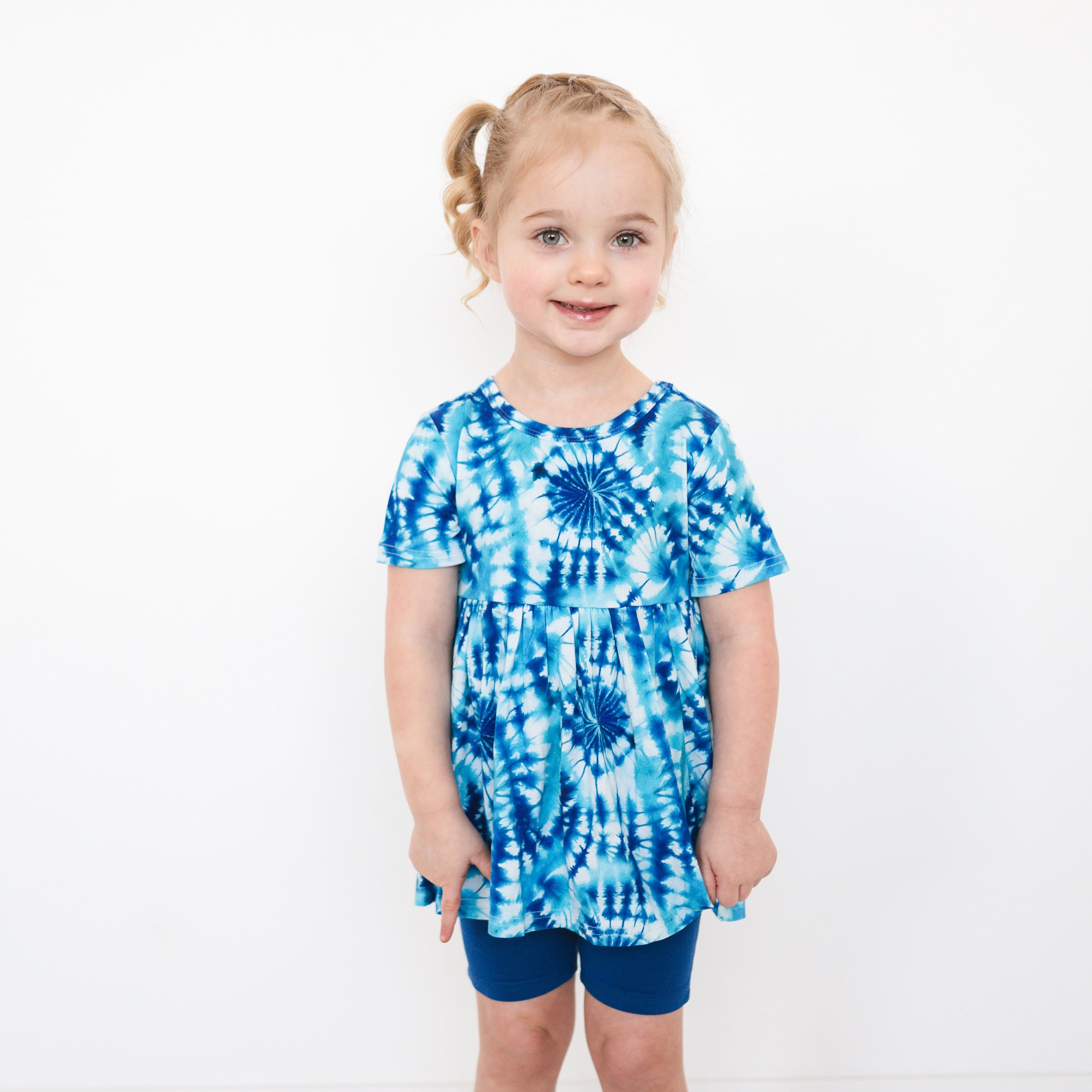 A young girl wears a blue tie-dye peplum top and navy shorts.