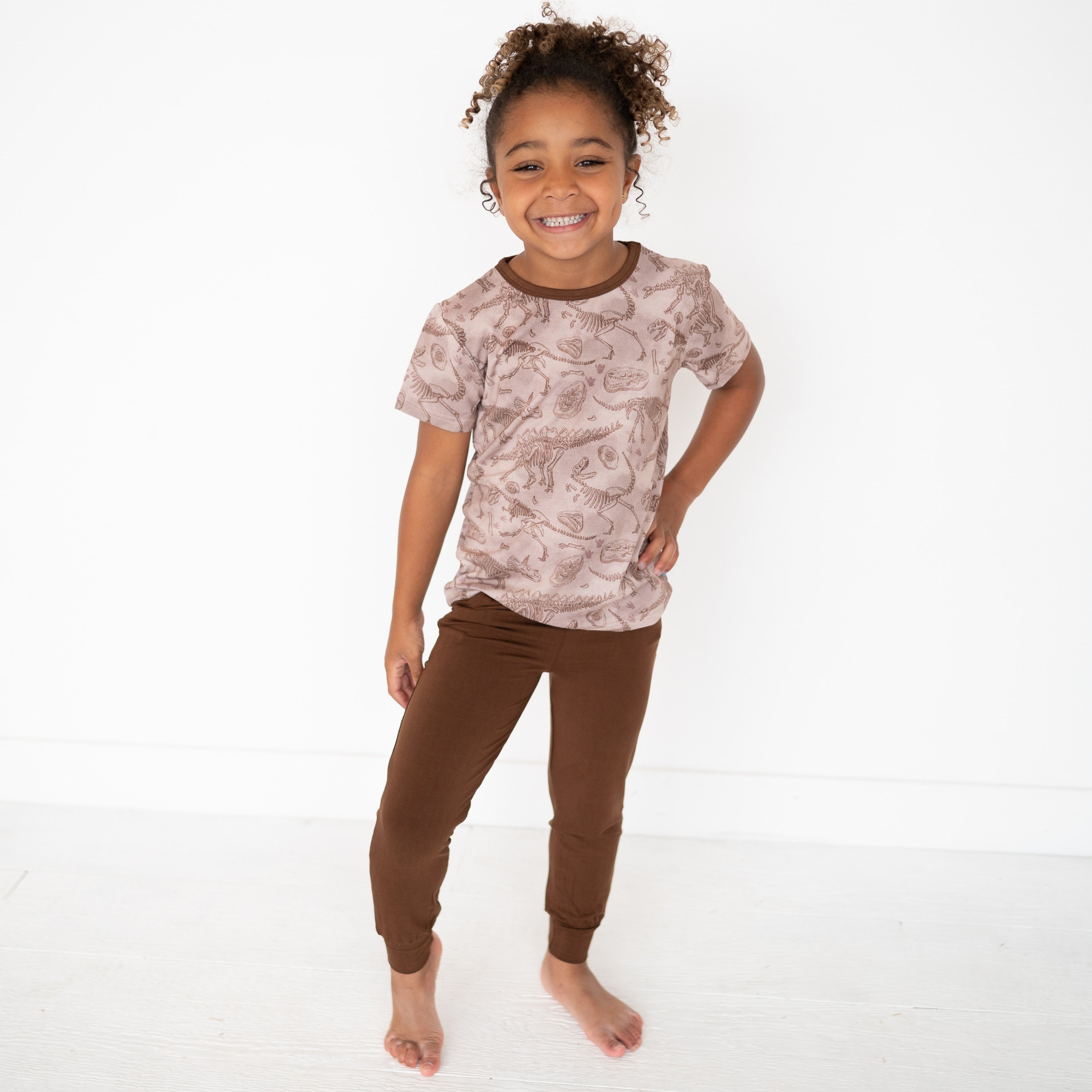 A young girl smiles in a brown dinosaur skeleton print t-shirt and brown joggers.