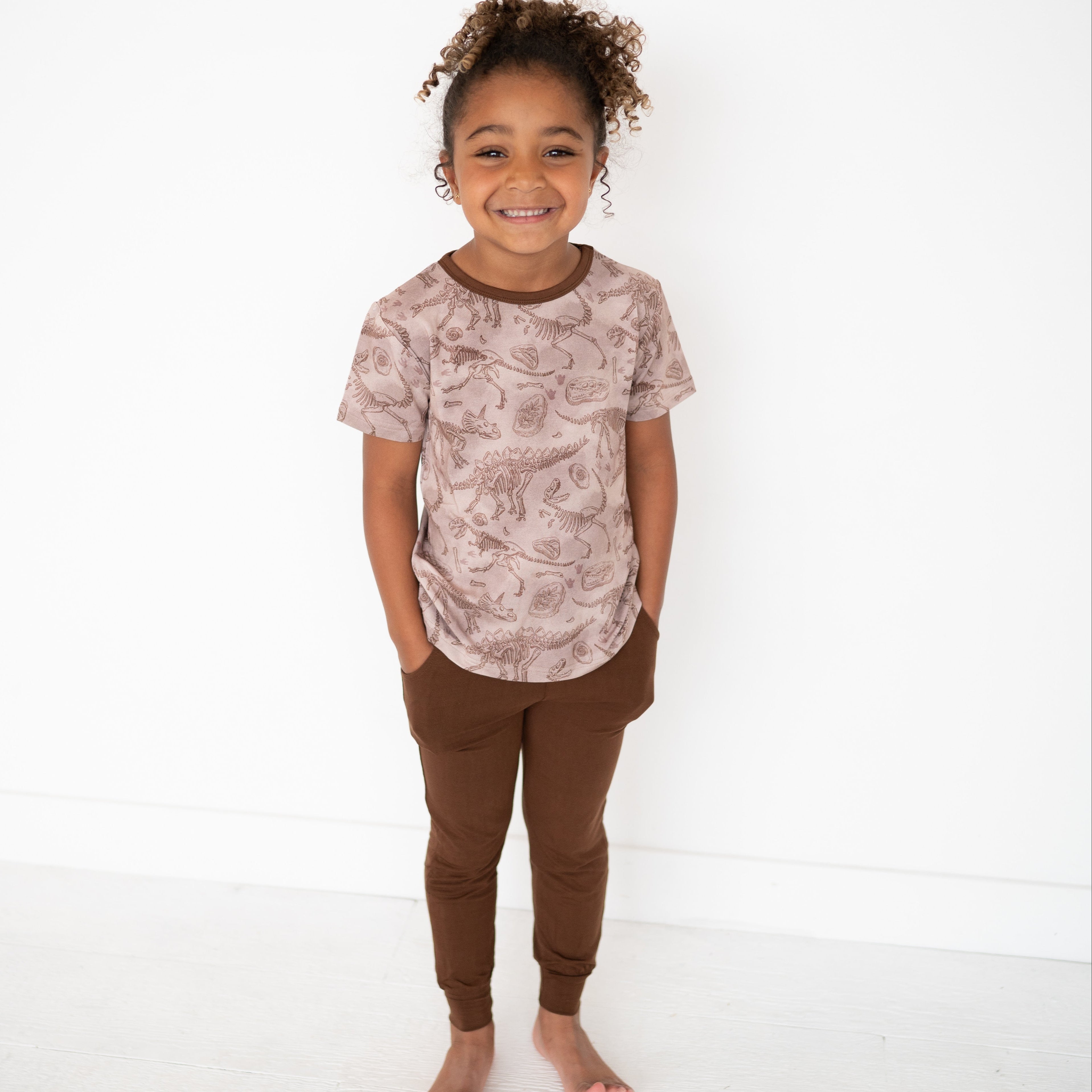A young girl smiles wearing a brown dinosaur print t-shirt and brown joggers.