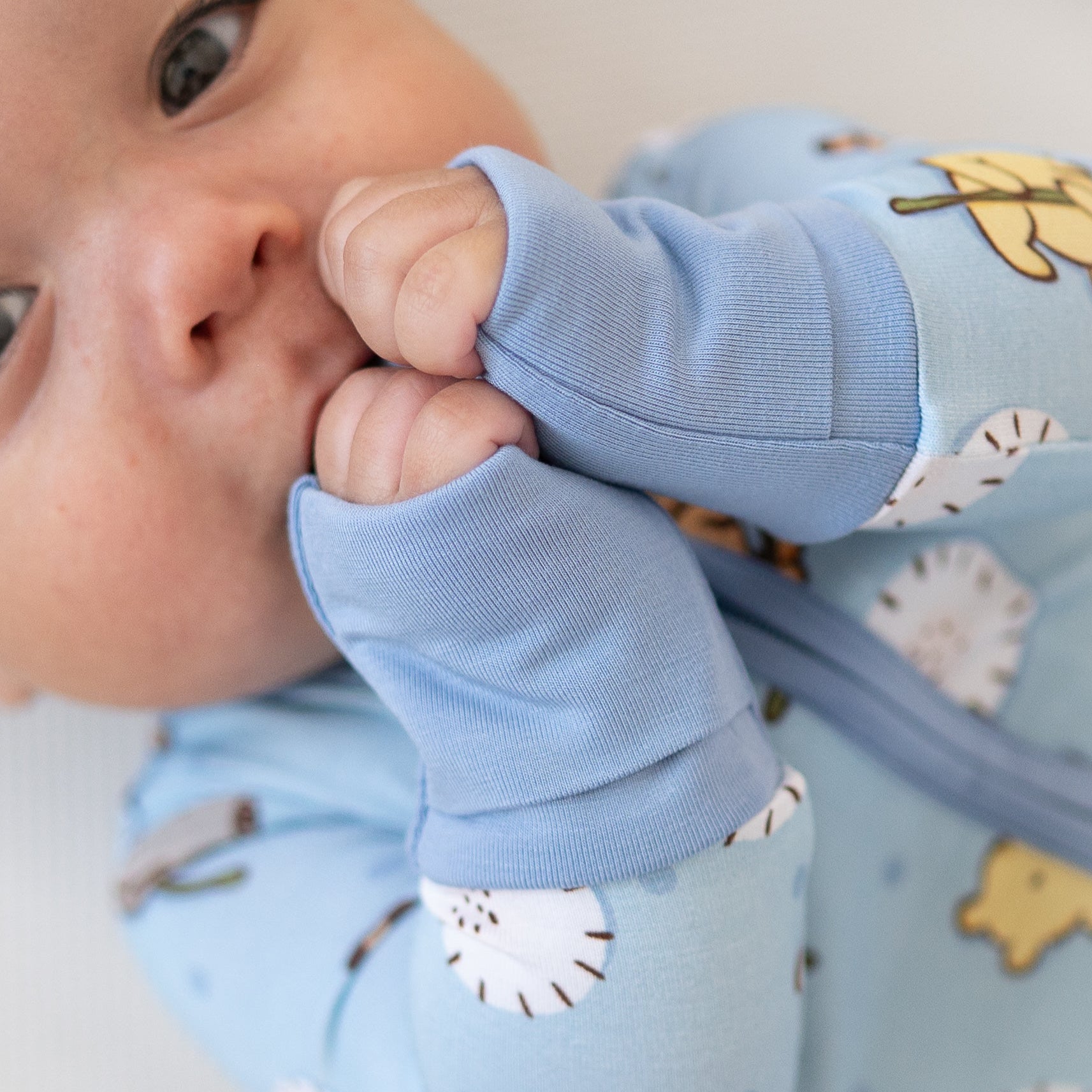 A baby in a light blue pajama set with a Winnie the Pooh print, sucking on their hands.