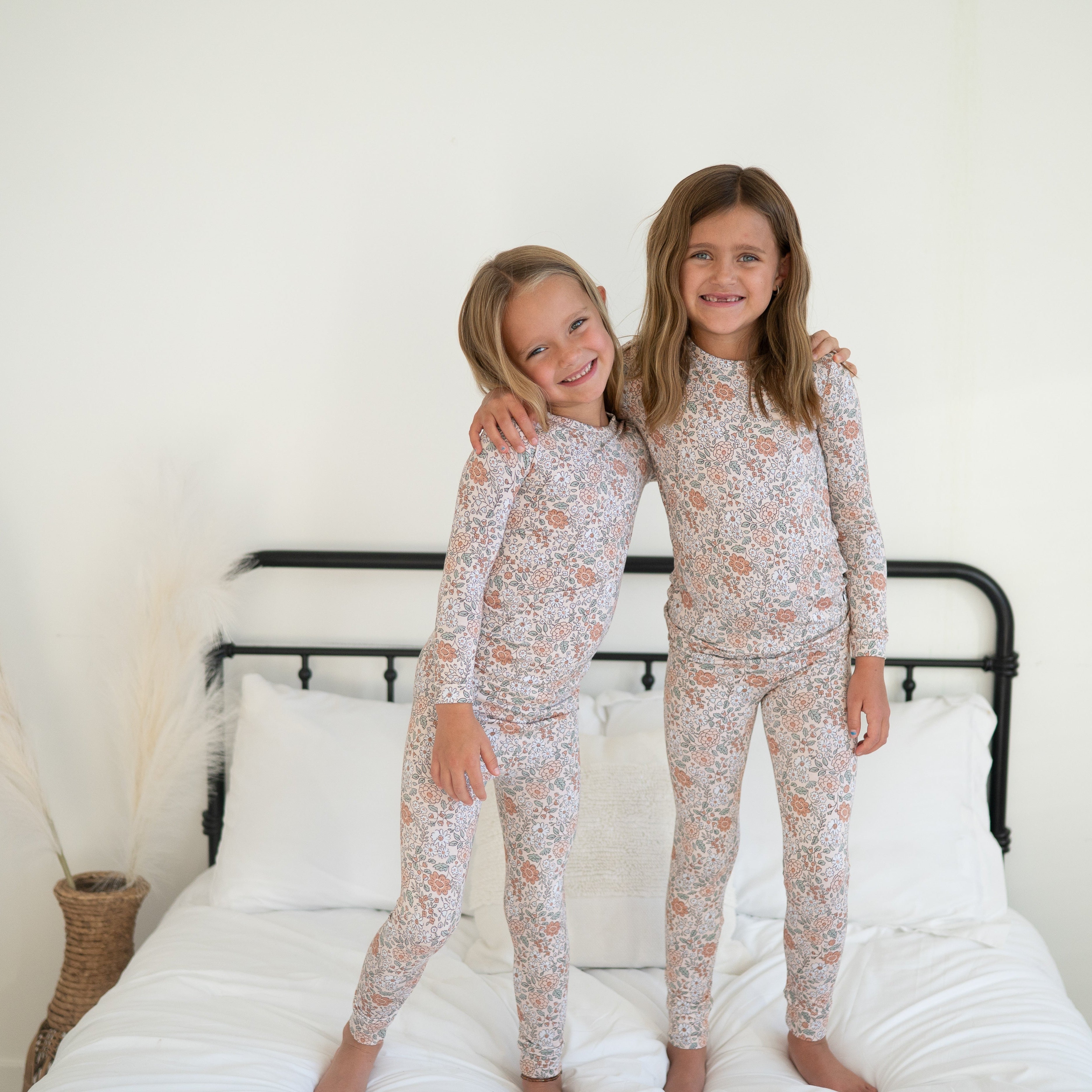 Two young girls hugging and smiling while wearing matching vintage floral long-sleeve pajama sets on a bed.