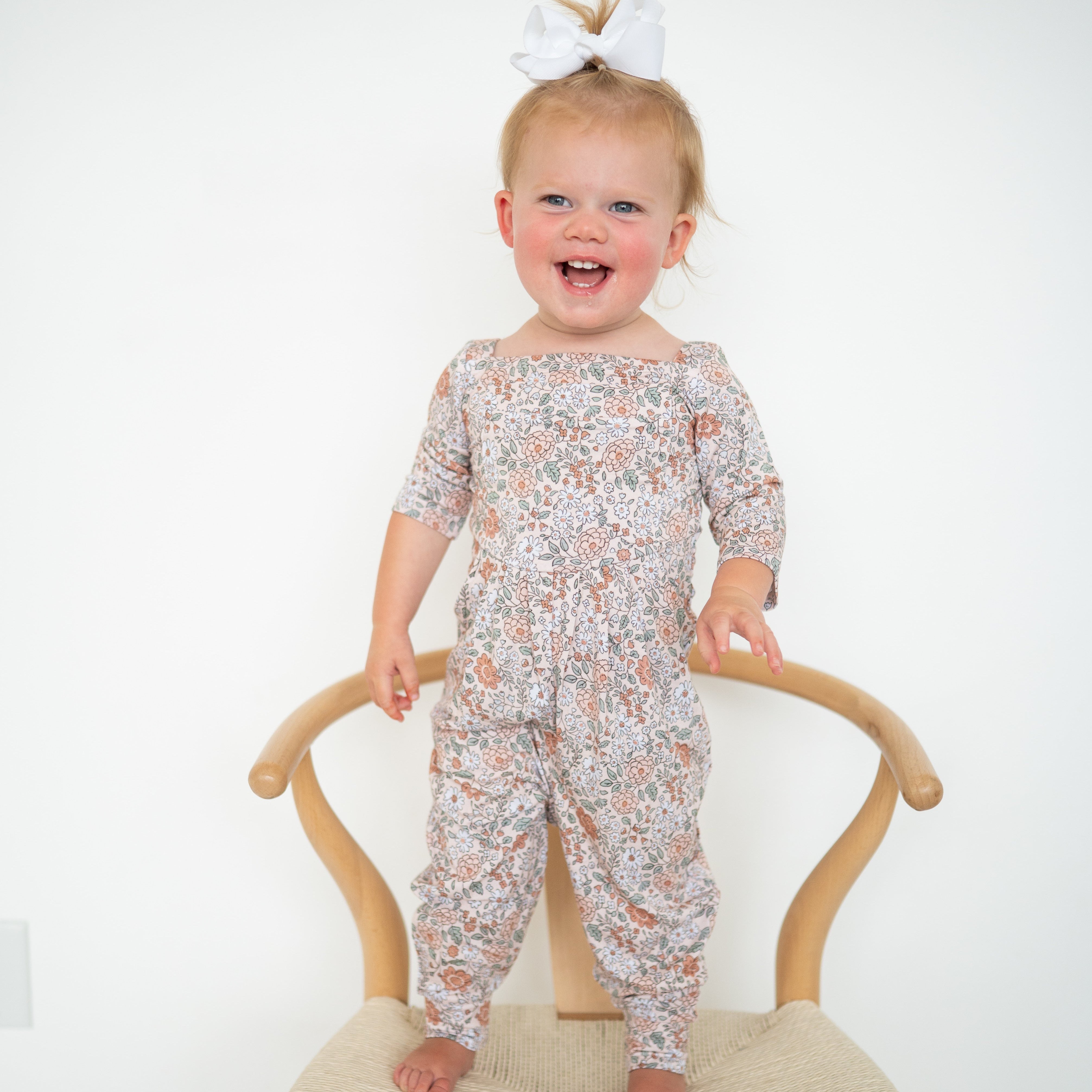 A toddler girl wearing a floral print romper stands on a wooden chair.