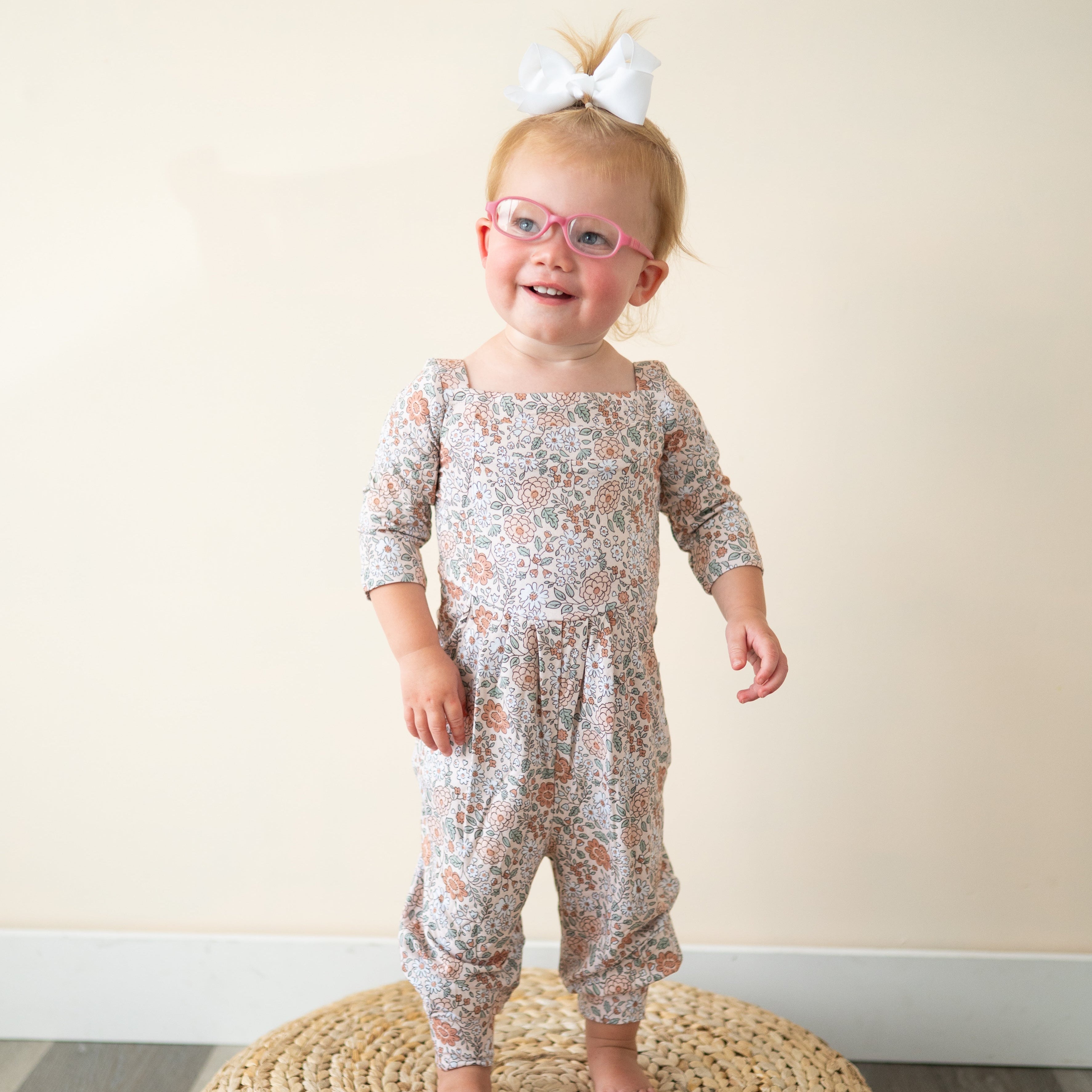 A toddler girl wears a vintage floral romper and pink glasses.