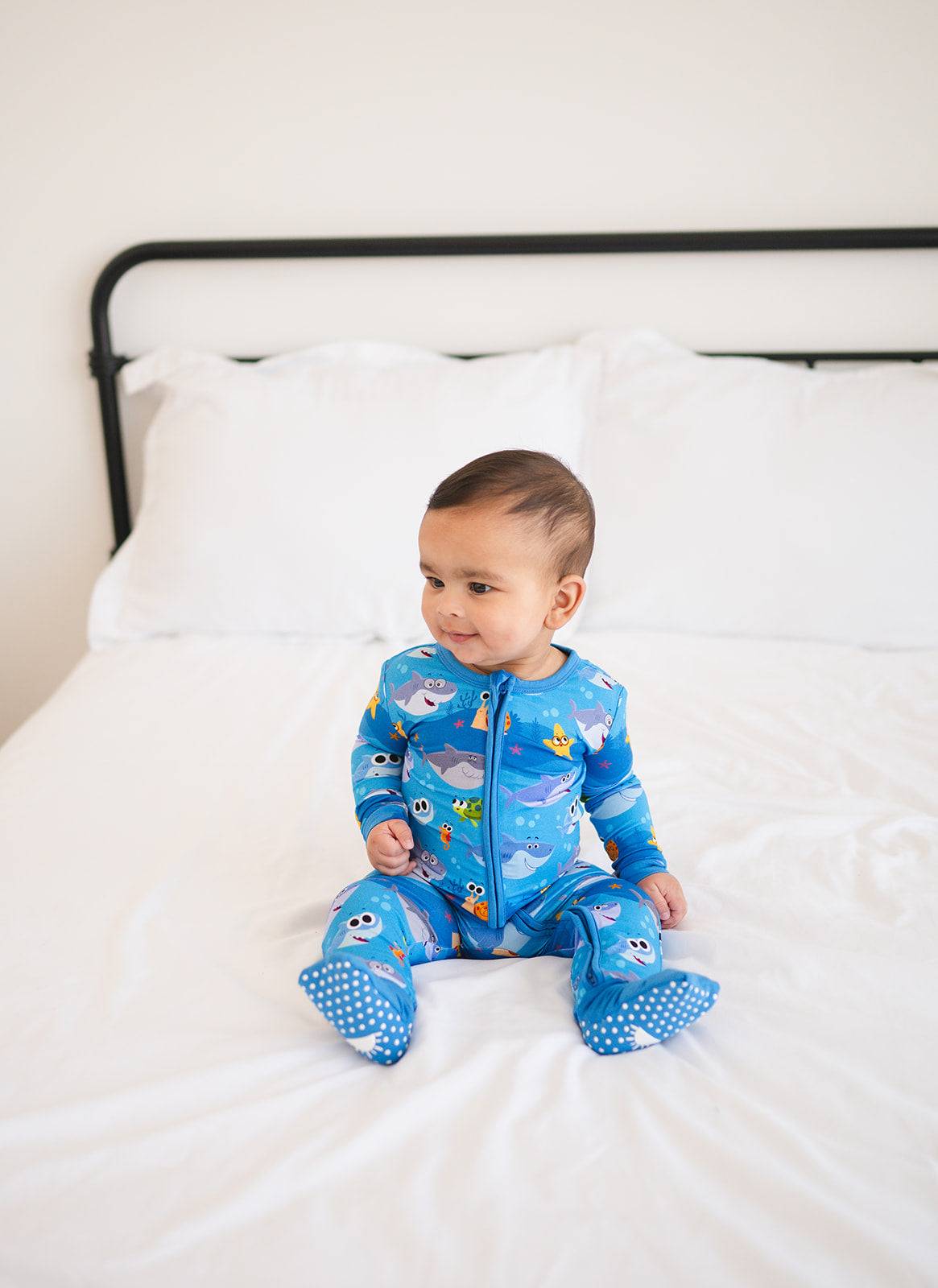 A baby boy sits on a bed wearing a blue footed pajama set with a Baby Shark print.