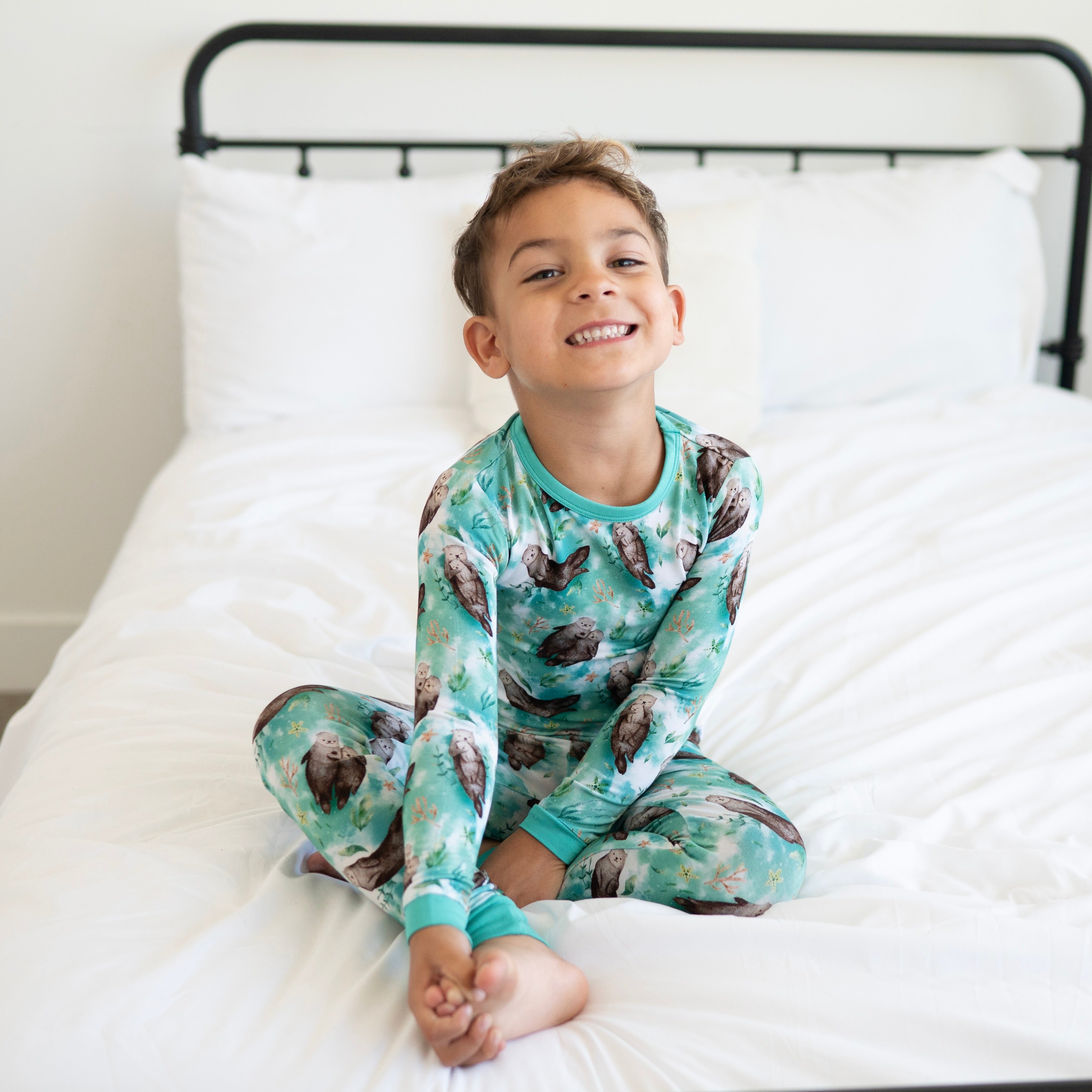 A young boy smiles while sitting on a bed wearing otter-print pajamas.