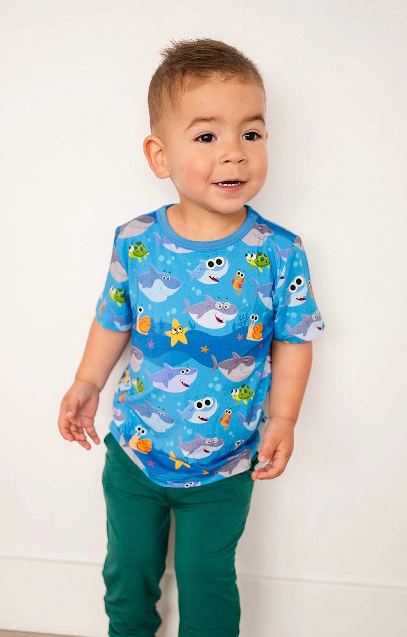 A young boy wears a blue t-shirt with a baby shark print and green pants.