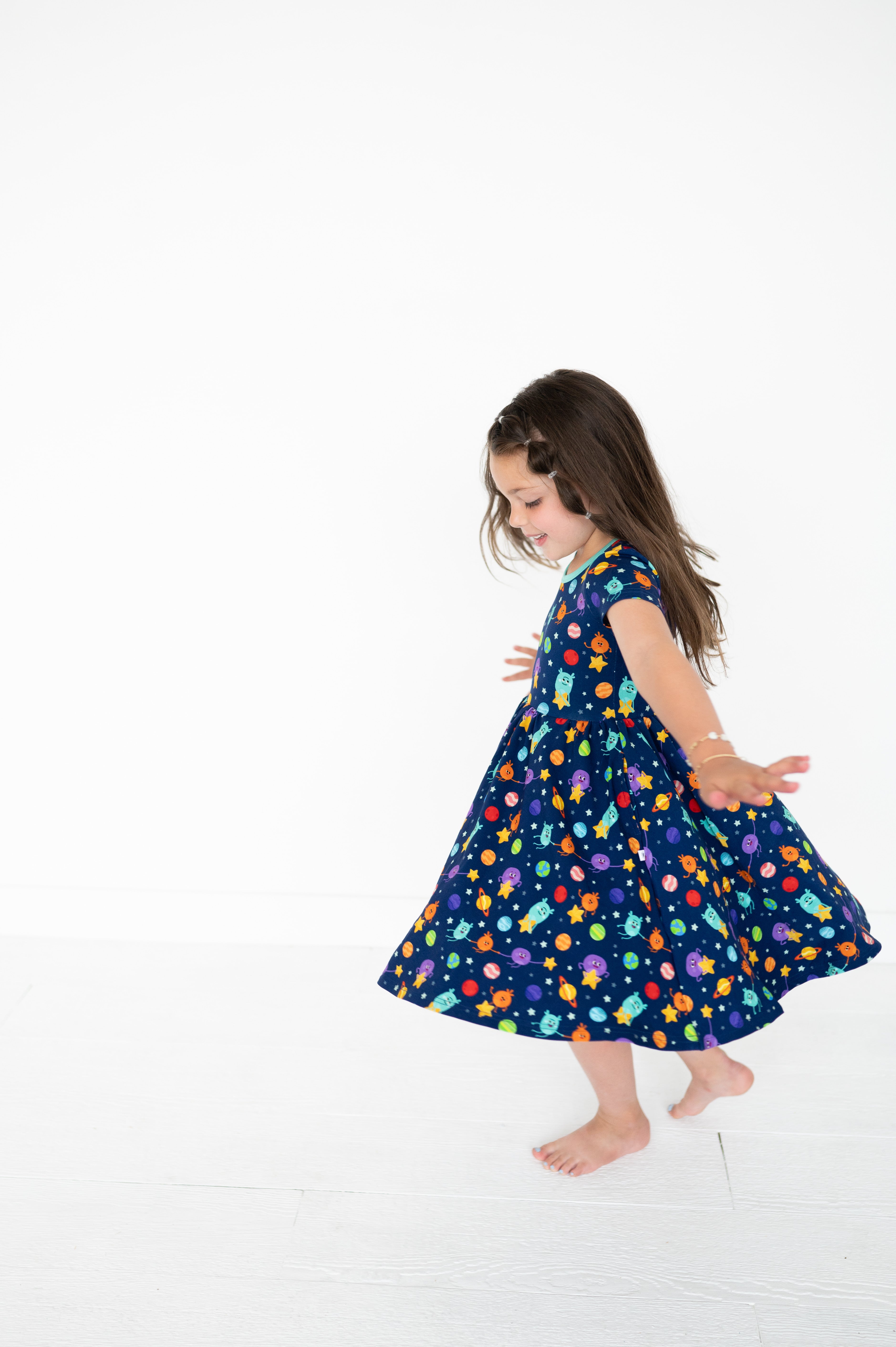 A young girl in a navy blue dress with a space-themed print twirls against a white background.