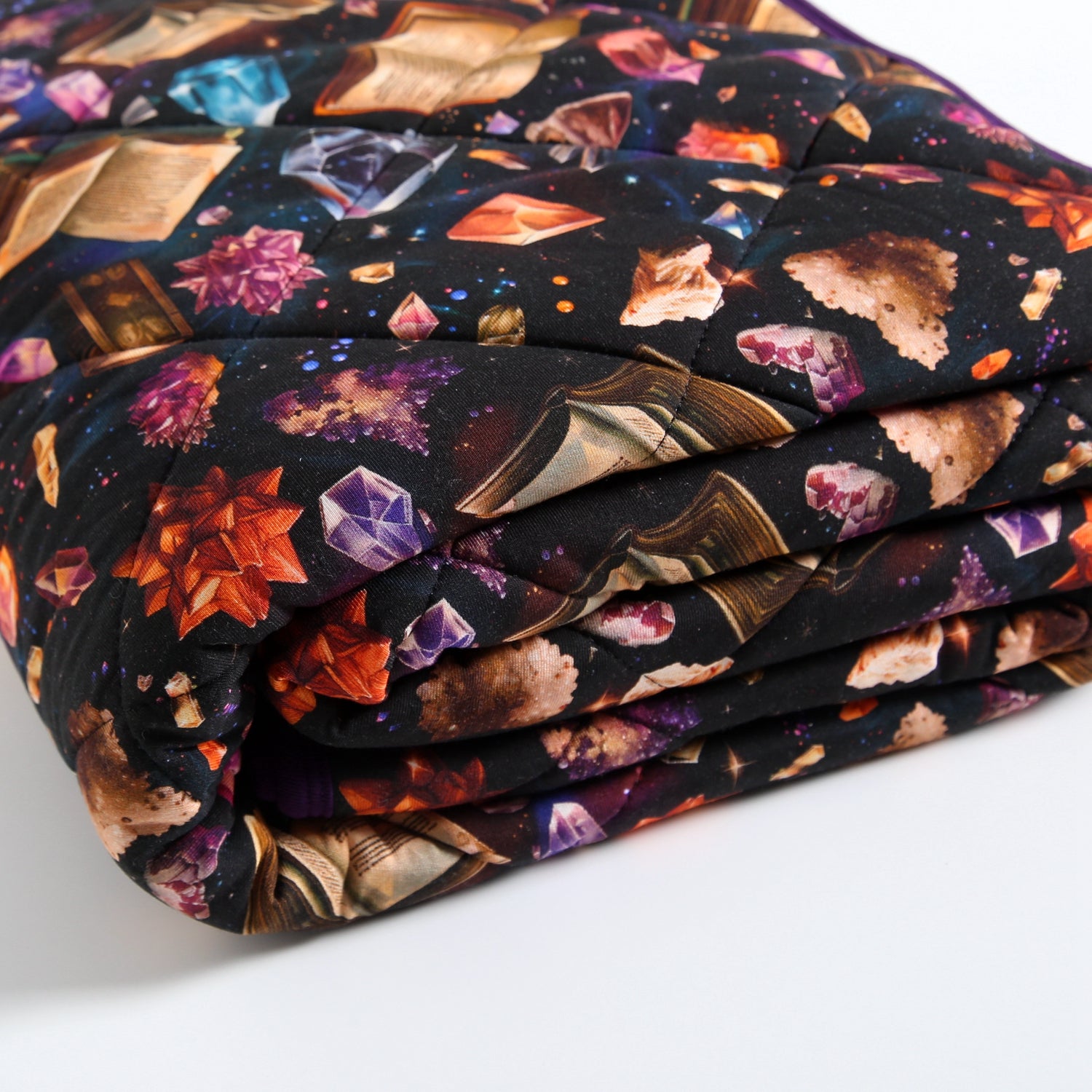 A folded quilted blanket with a black background featuring a print of open books and colorful crystals.