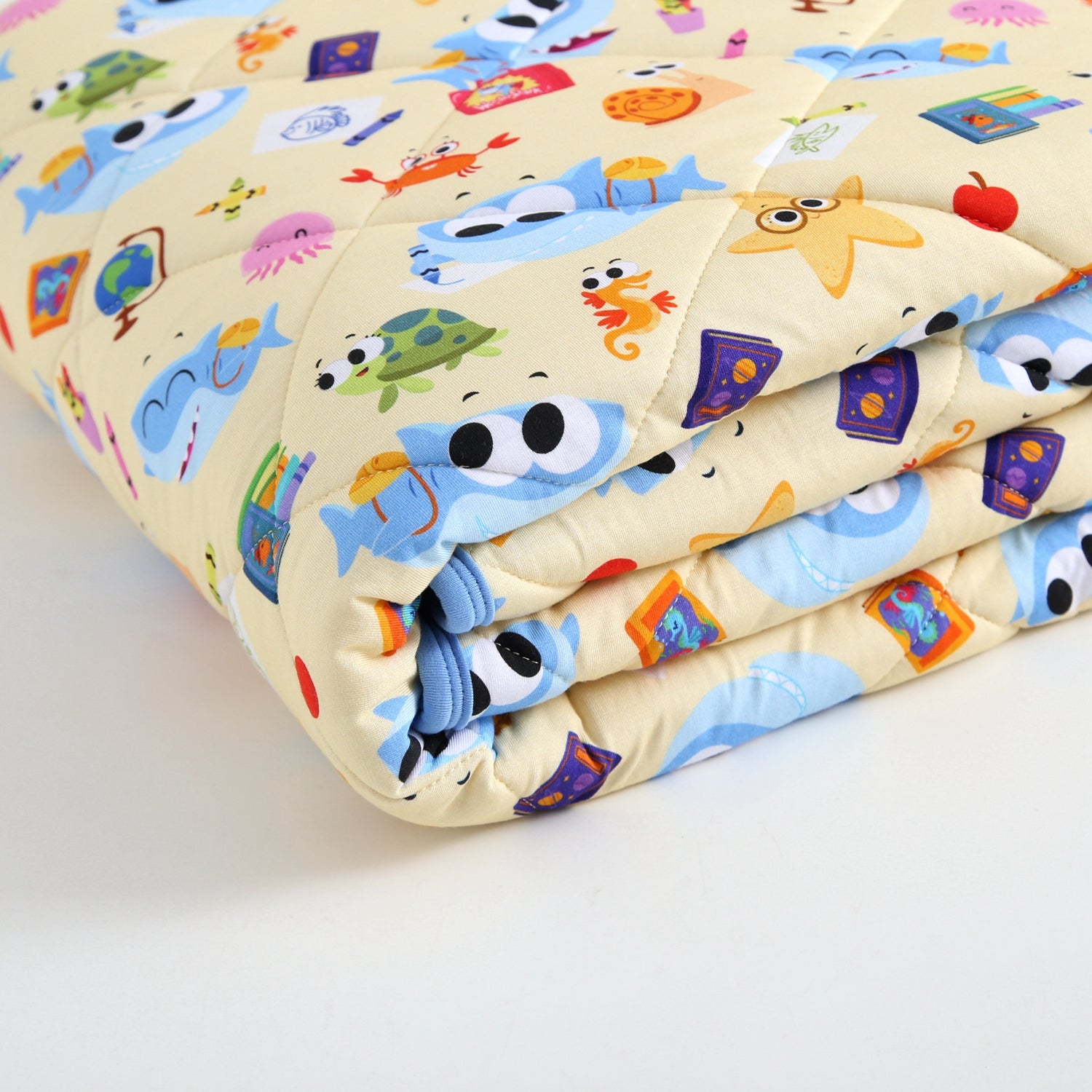A folded light yellow quilted blanket with a pattern of cartoon sea creatures and school supplies.