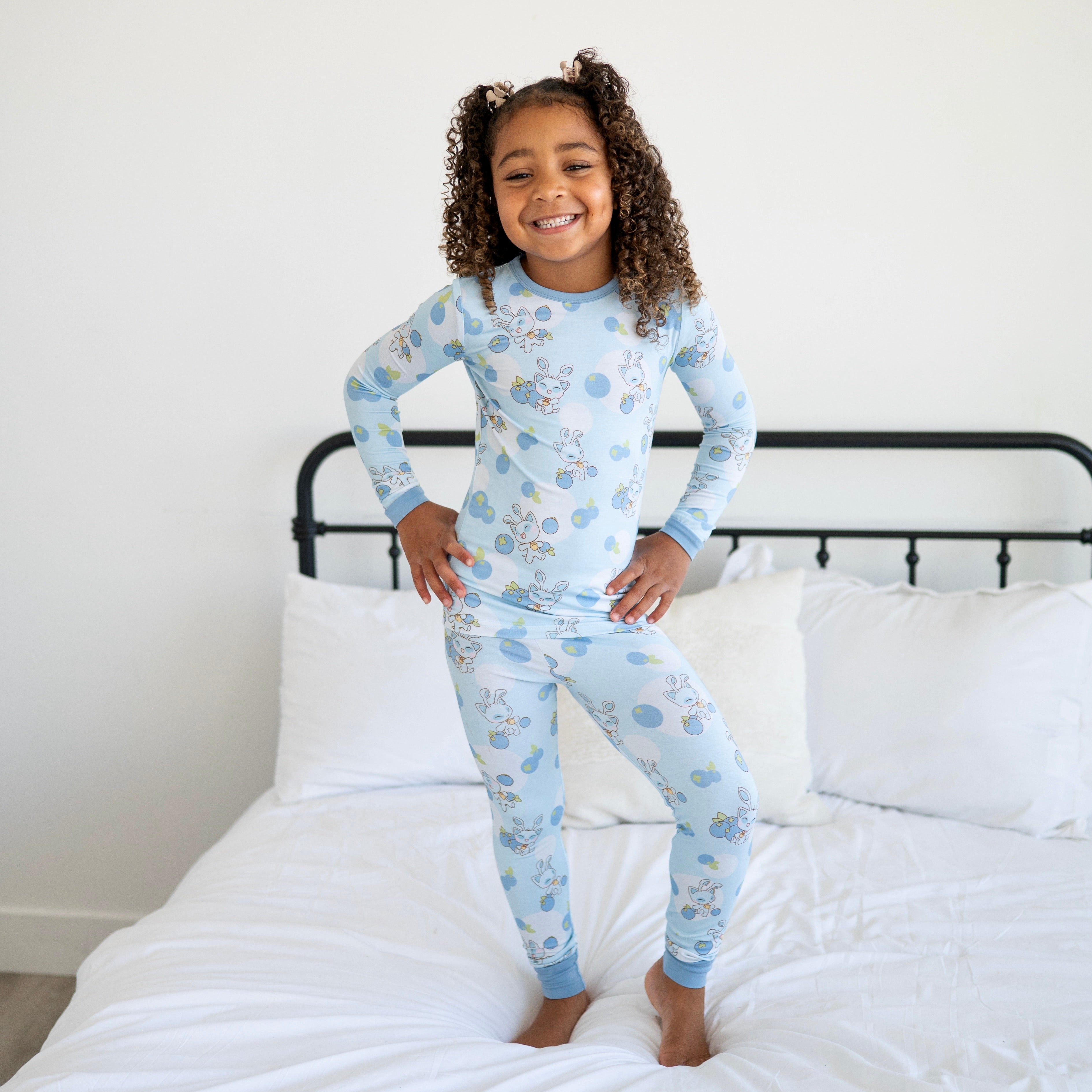A young girl in blue pajamas with a blueberry and bunny pattern stands on a bed.