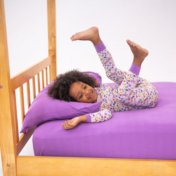 A young girl in floral pajamas plays on a twin bed with lavender sheets.