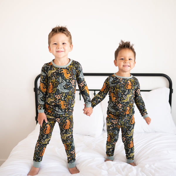 Two young boys in matching dark pajamas with a jungle print stand on a bed.