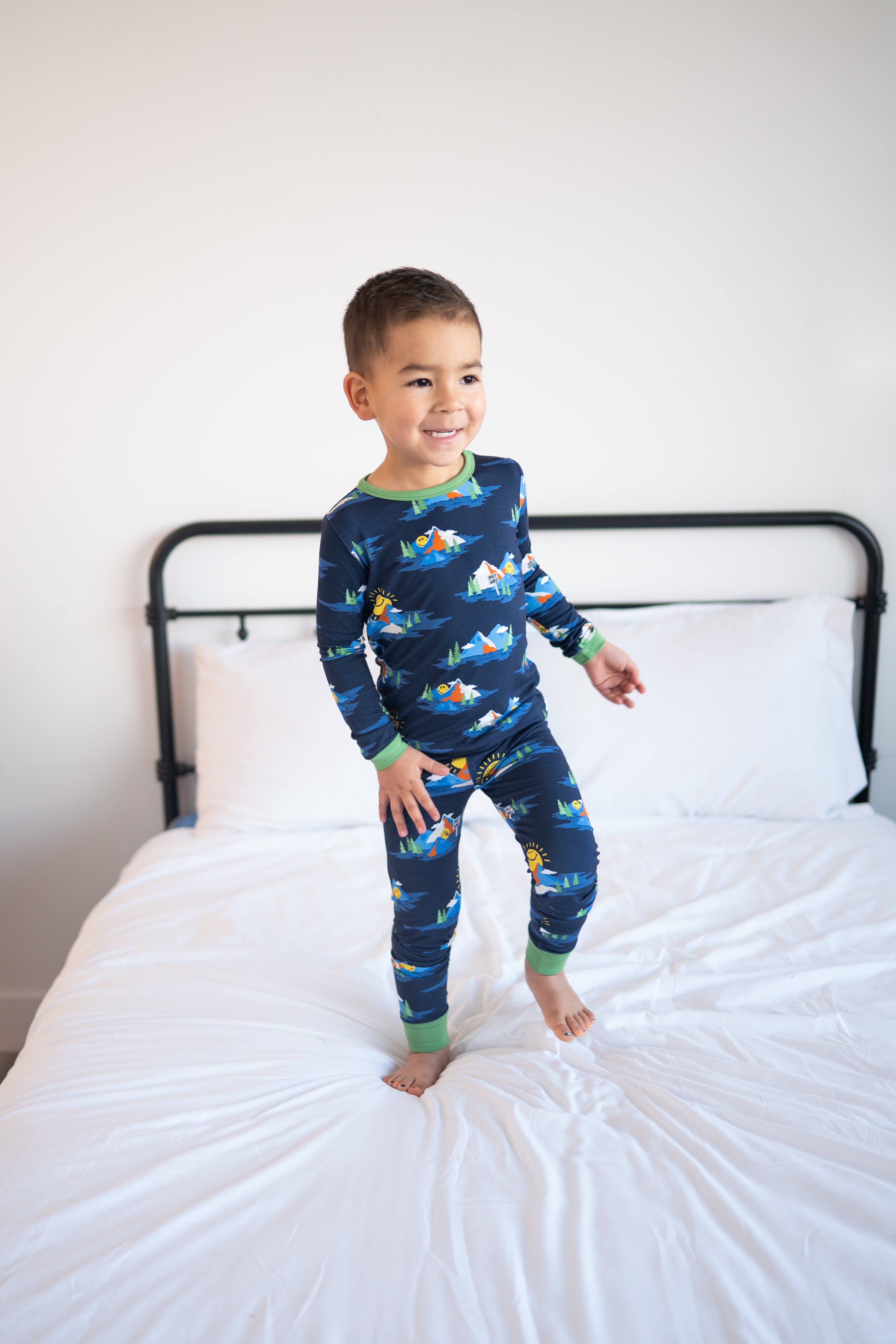 A young boy in navy blue pajamas with a mountain and smiley face print stands on a white bed.